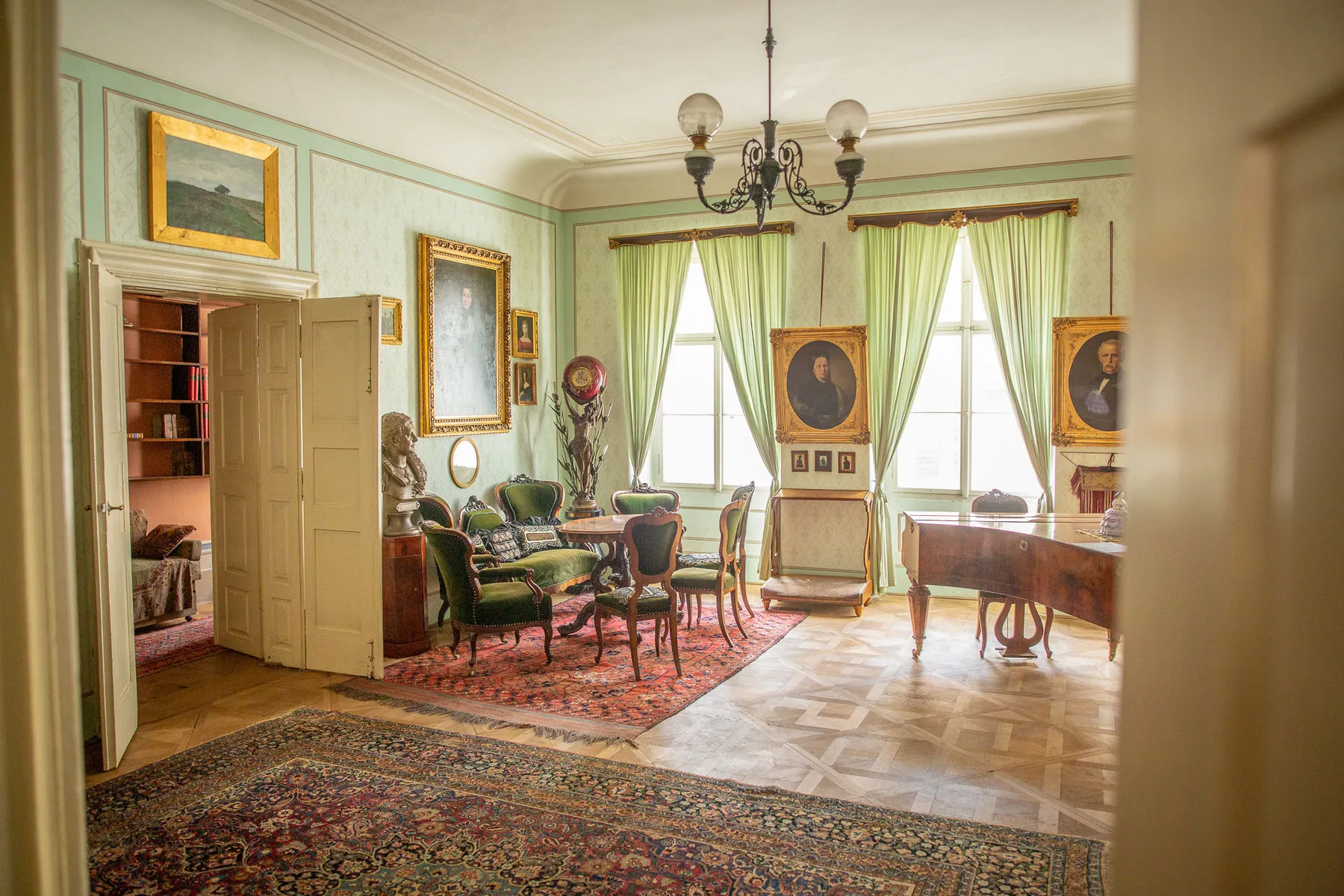 Interieur van een historische kamer met groene muren, houten meubels, ingelijste portretten en een kroonluchter.