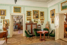 Interieur van een historische kamer met lichtgroene muren, ingelijste kunstwerken en meubilair uit die periode, waaronder groene stoelen en een houten tafel.