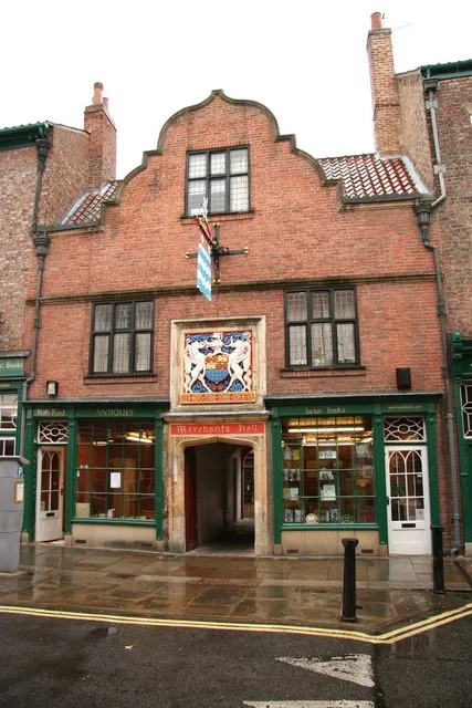 Brick facade of the Merchant Adventurers' Hall with a curved gable, coat of arms above the entrance, and green-framed shop windows on a wet street