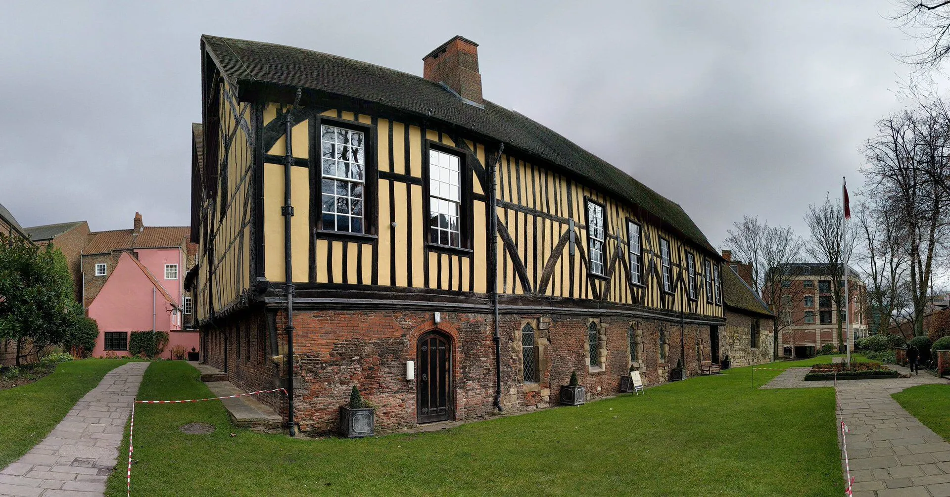 Timber-framed medieval guildhall with yellow and black half-timbered upper story on a red brick base, set on a green lawn under an overcast sky