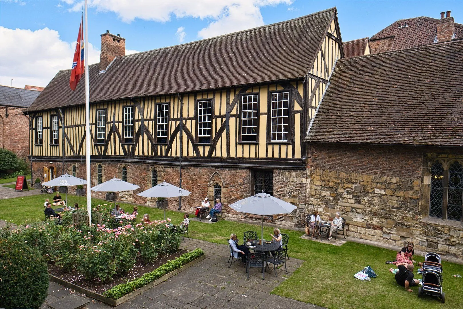 Exterior view of the Merchant Adventurers' Hall, a large timber-framed building with a courtyard garden featuring outdoor seating under white umbrellas and a flagpole.