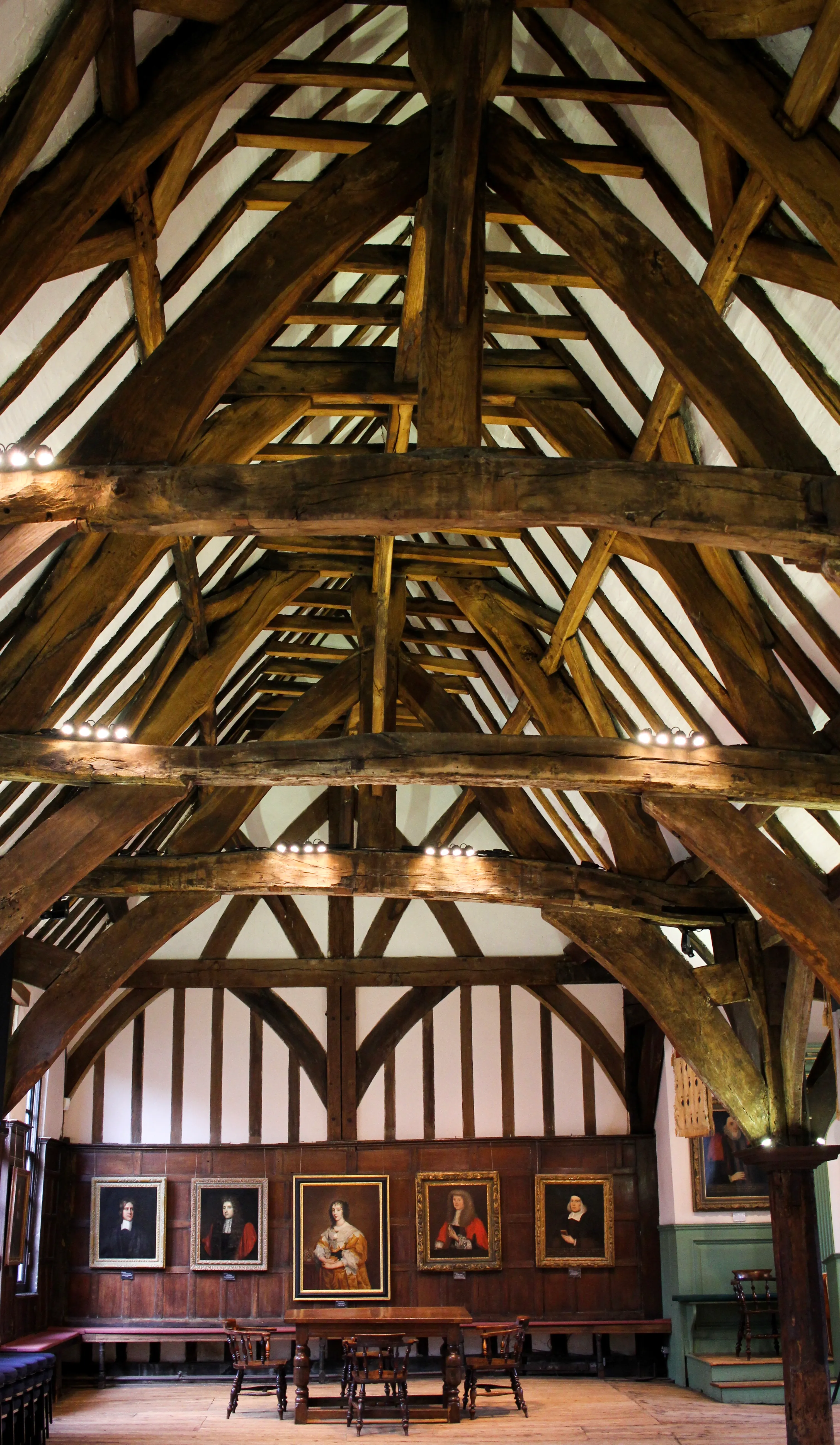 Medieval timber-framed ceiling with massive wooden beams and trusses above a great hall with portrait paintings in gilded frames on dark wood-paneled walls and a long table with chairs