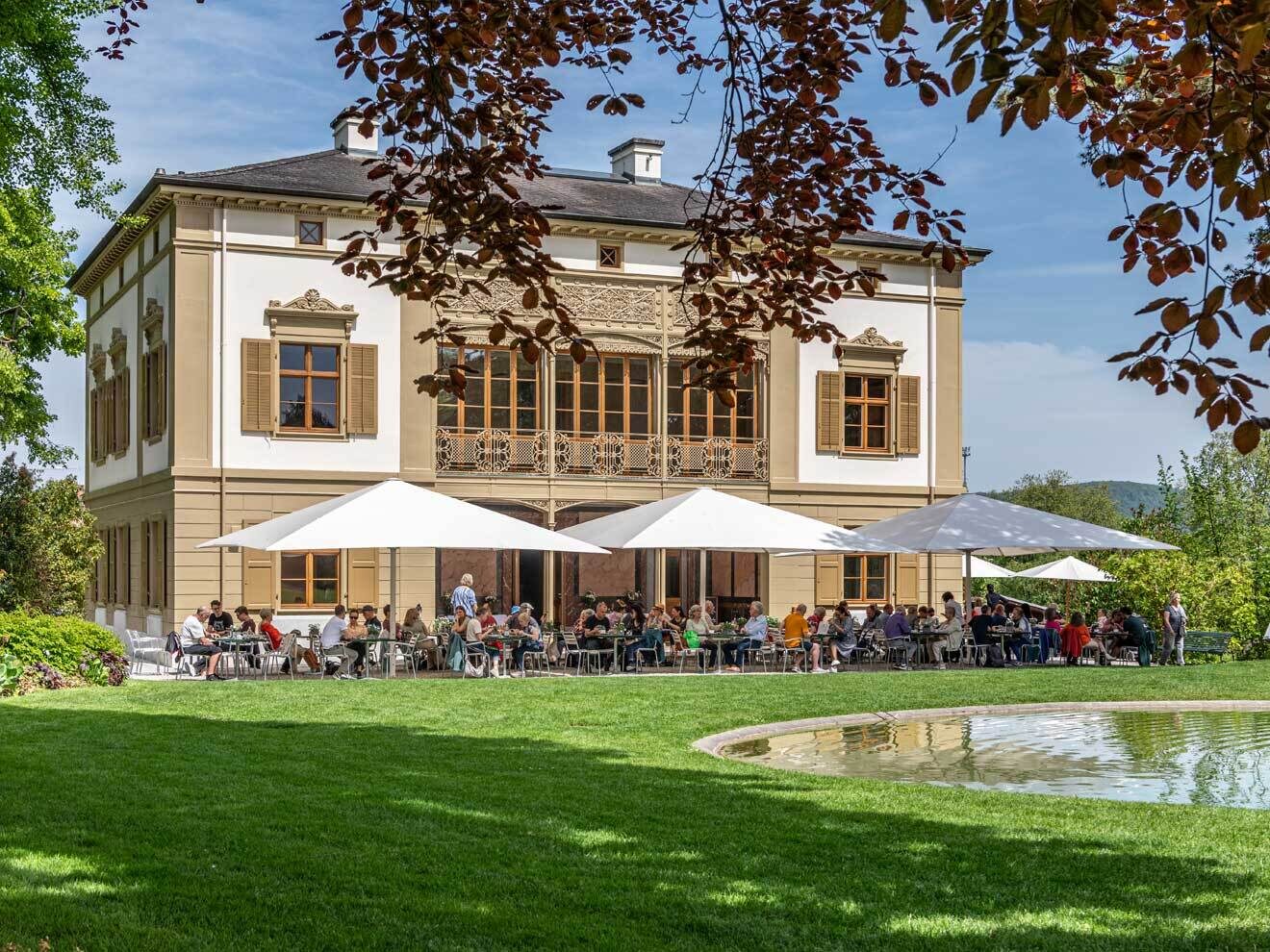Historisch villa-gebouw in de Merian Gärten met terras met buiten-café onder witte parasols, met een groen gazon en vijver op de voorgrond
