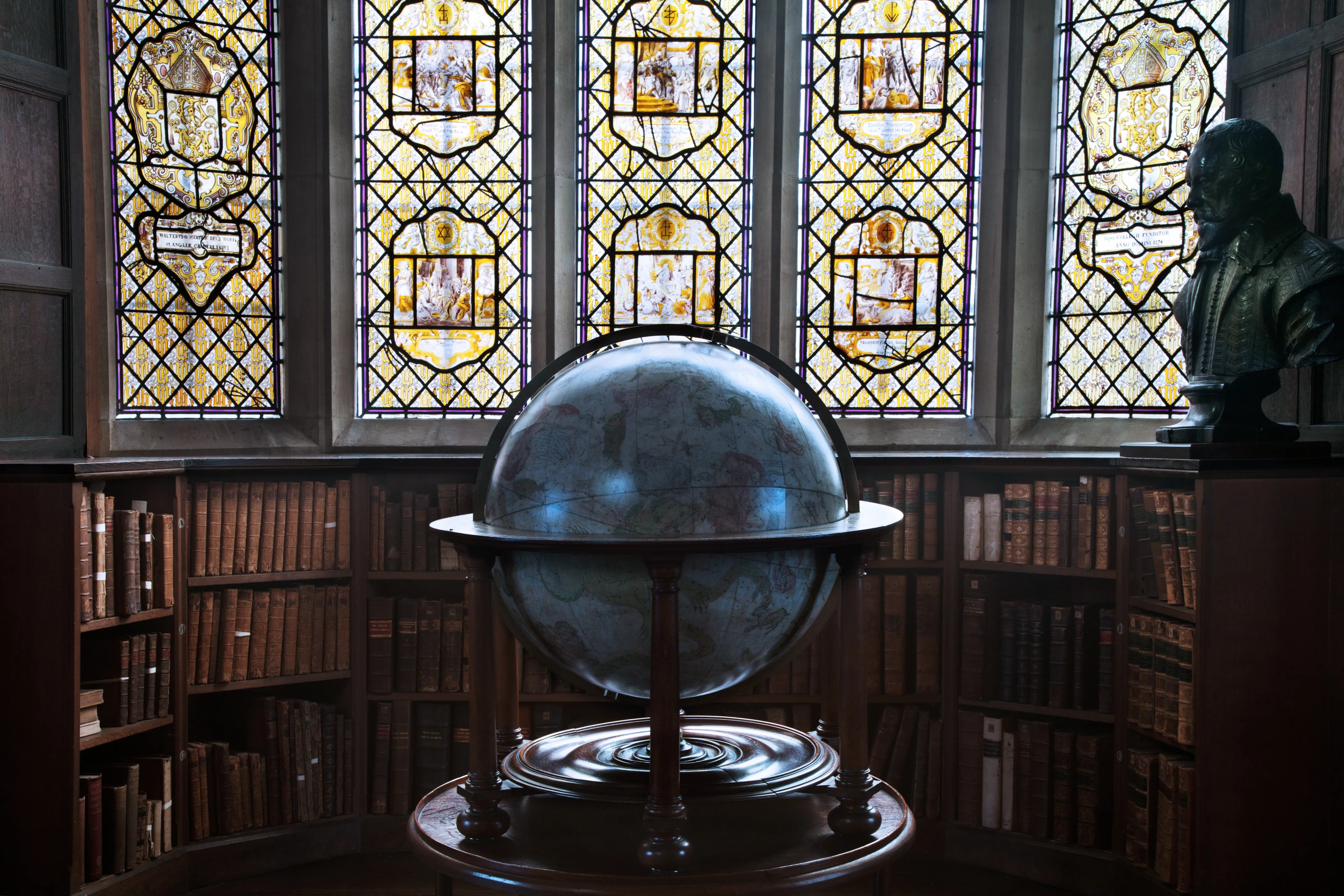 Un gran globo terráqueo antiguo sobre un soporte de madera, situado frente a altos ventanales de vitrales en una biblioteca histórica llena de estanterías y un busto.