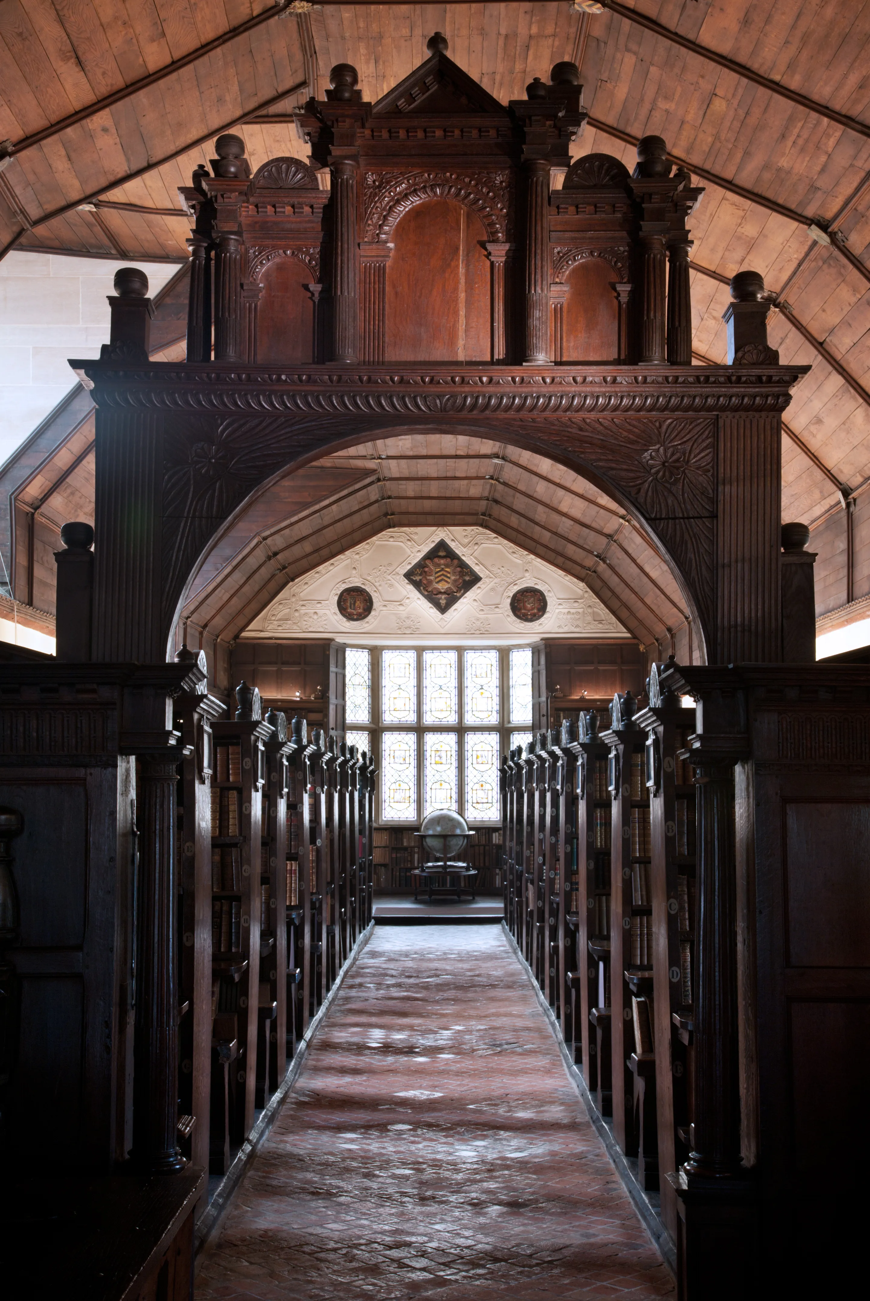 Vista interior de la histórica biblioteca de Merton College que muestra hileras de estanterías de madera oscura a ambos lados de un pasillo central bajo un arco de madera tallada ornamentado y un techo abovedado.