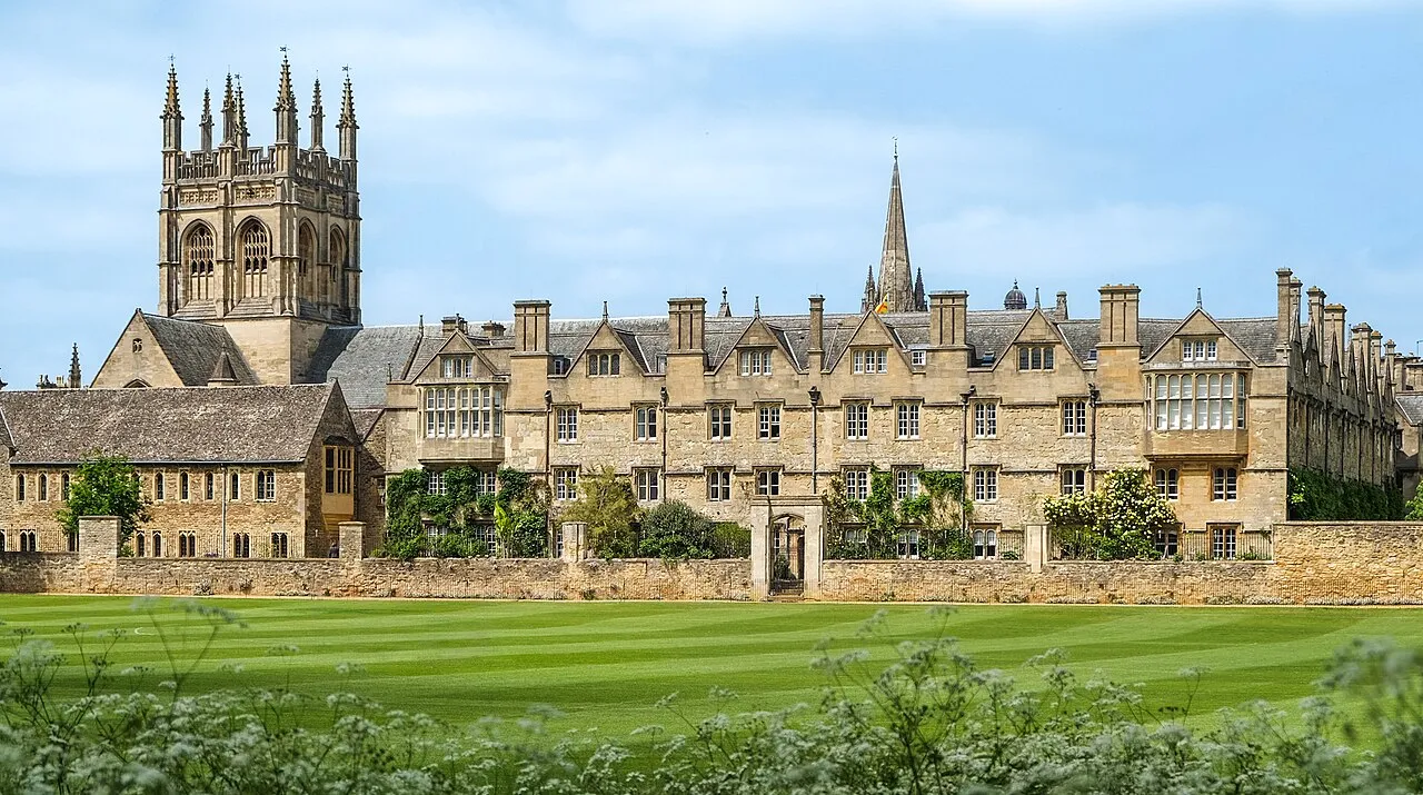 Vista exterior amplia del Merton College de Oxford que muestra históricos edificios de piedra, la torre de la capilla y un gran césped bajo un cielo azul.