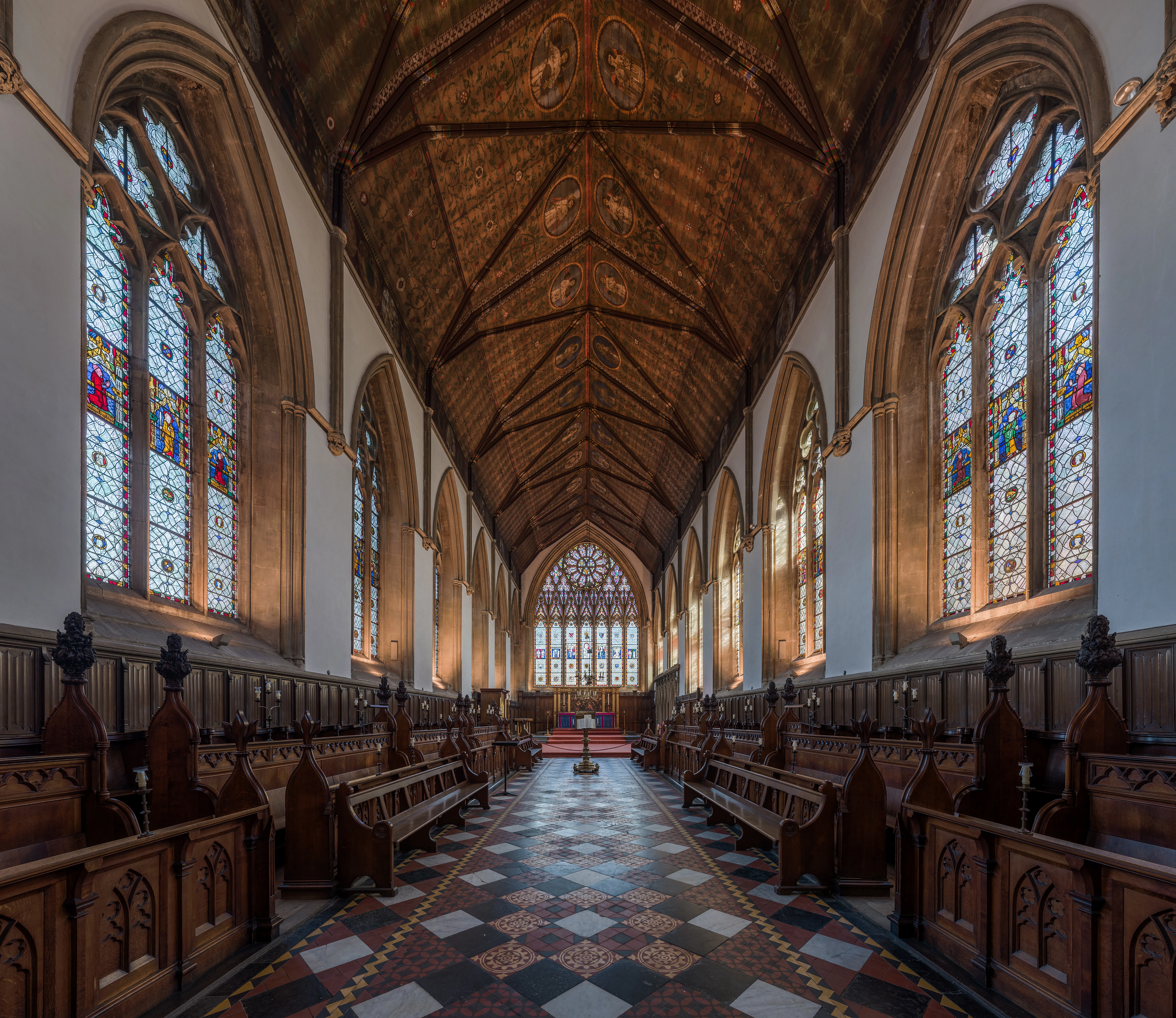 Interior de la capilla del Merton College que muestra hileras de sillería de coro de madera, altos vitrales, un techo de madera ornamentado y un suelo de baldosas a cuadros que conduce al altar.