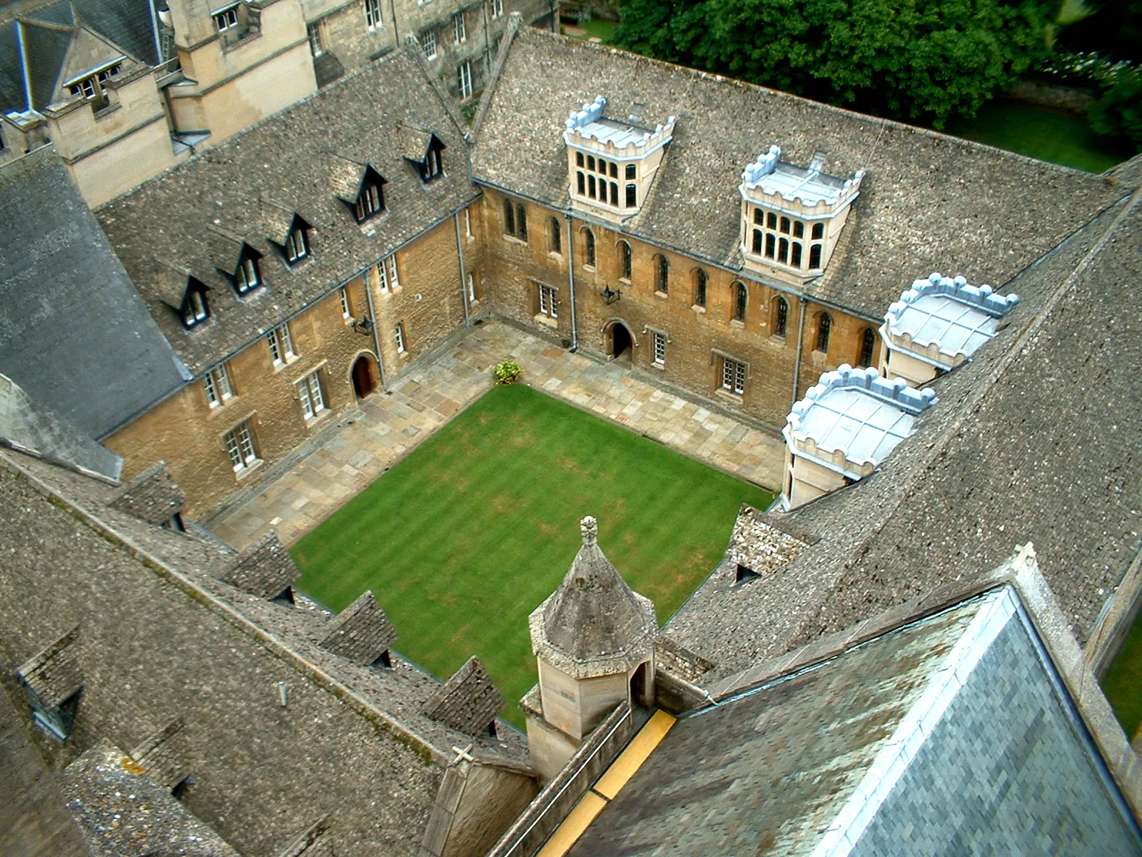 Vista aérea de un histórico patio de piedra con un césped central, rodeado de edificios con tejados de pizarra y buhardillas en Merton College.