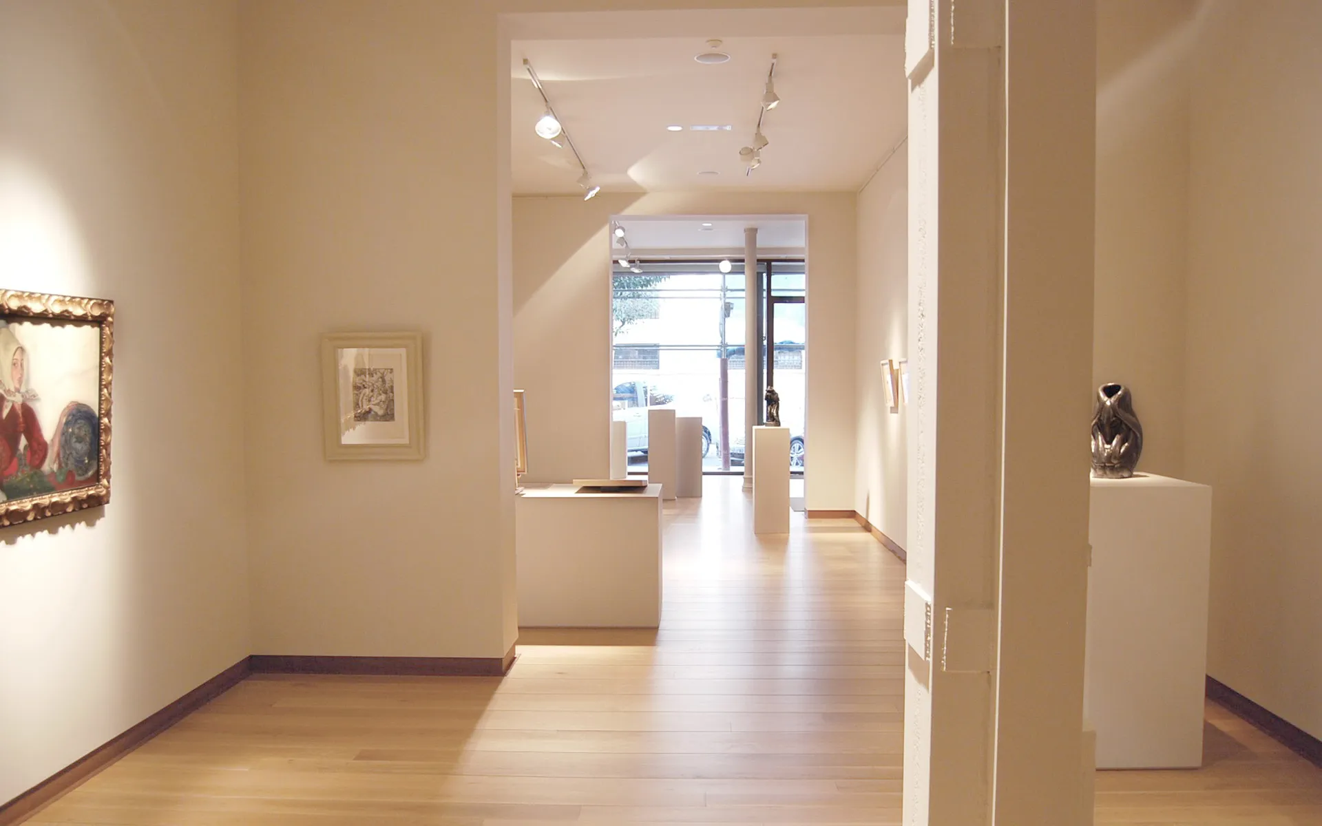 Licht interieur van een galerie met witte muren, houten vloeren, railverlichting, ingelijste schilderijen aan de muren en een sculptuur op een witte sokkel.