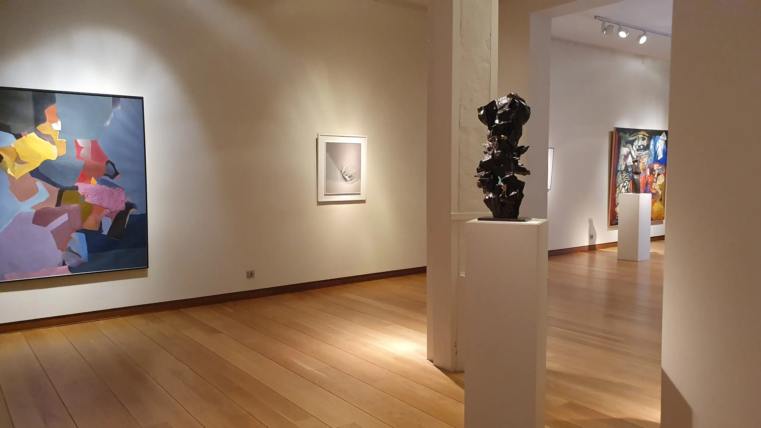 Interieur van een kunstgalerie met houten vloeren, witte muren, een groot kleurrijk schilderij aan de linkerkant, een klein ingelijst kunstwerk in het midden en een donker beeldhouwwerk op een witte sokkel.