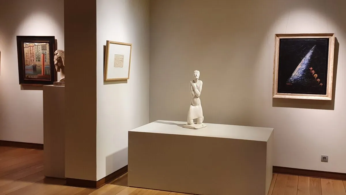 Een wit beeldhouwwerk van een staande figuur op een rechthoekig voetstuk in een galerieruimte, omringd door ingelijste schilderijen aan witte muren.