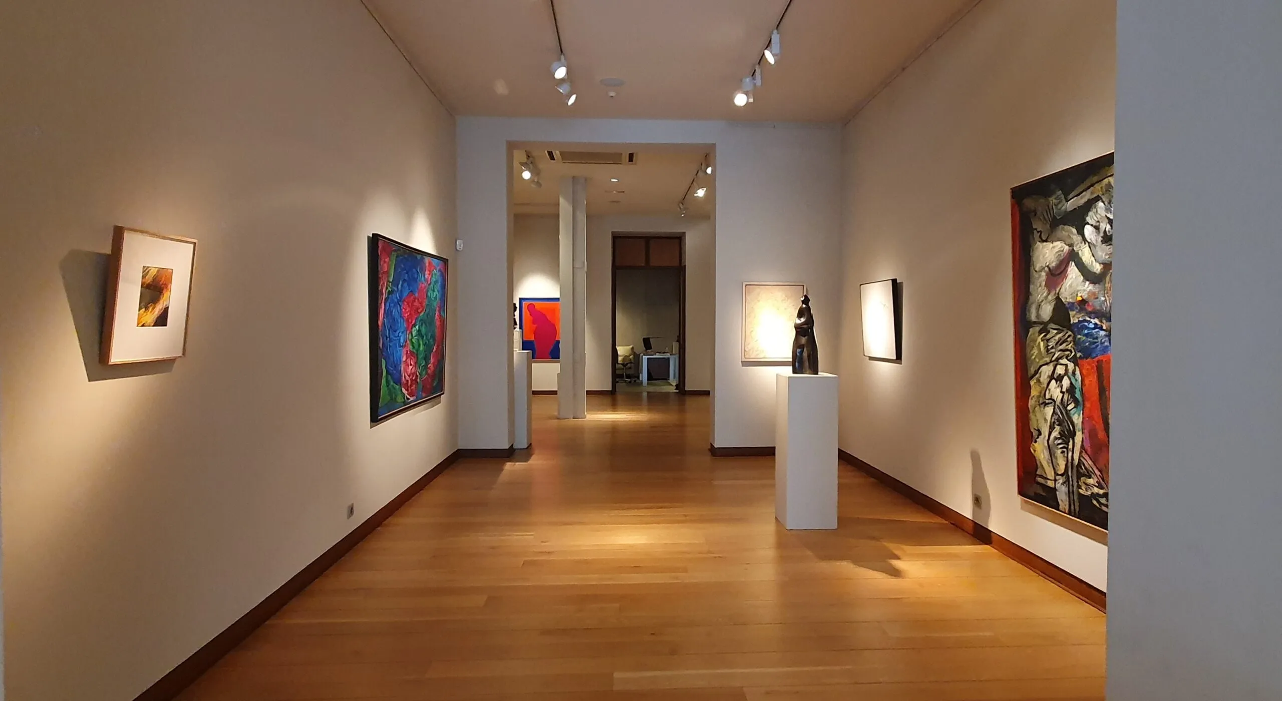 Lange galerijhal met houten vloeren, witte muren, railverlichting en diverse schilderijen en een sculptuur opgesteld.