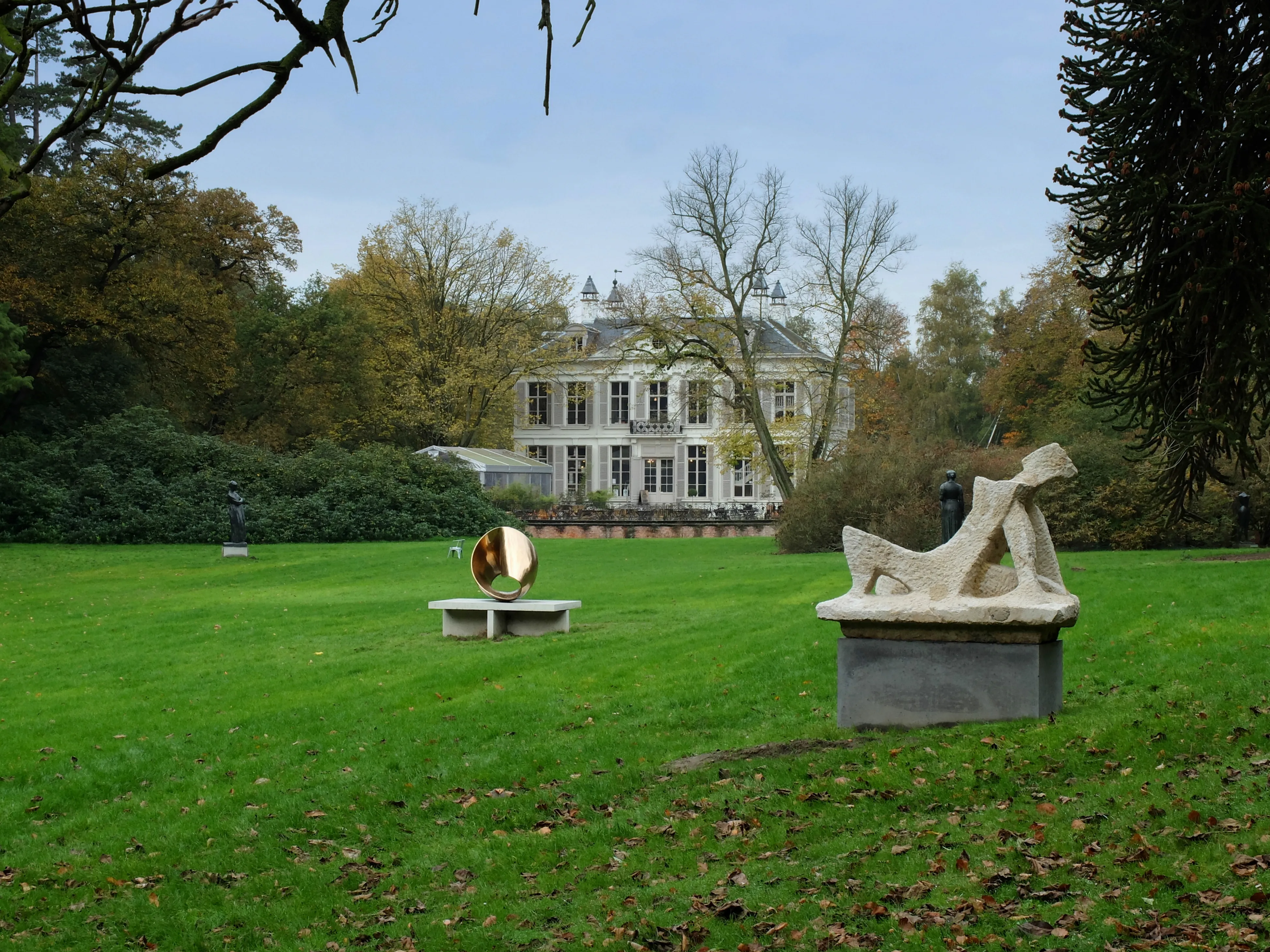 Césped verde con esculturas modernas, incluyendo una figura reclinada sobre un pedestal y una escultura circular, con una histórica villa blanca de fondo rodeada de árboles de otoño