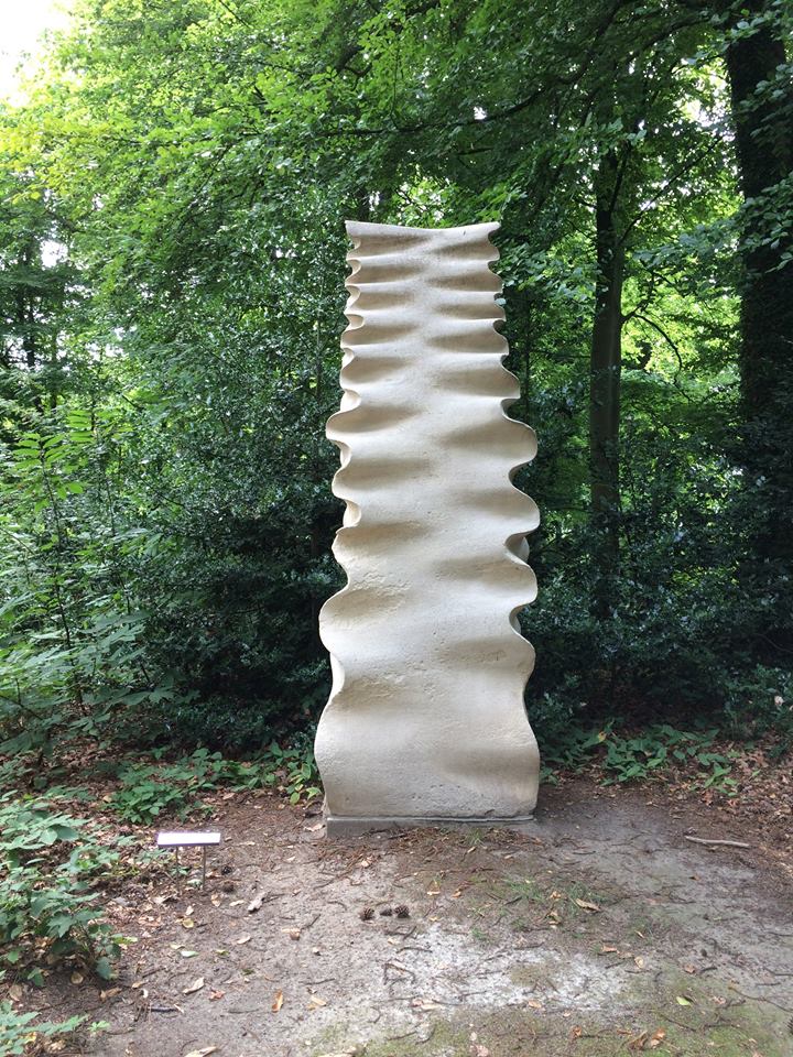 Escultura de piedra alta, blanca y ondulada, situada en un camino de tierra rodeado de densos árboles verdes en un parque arbolado.