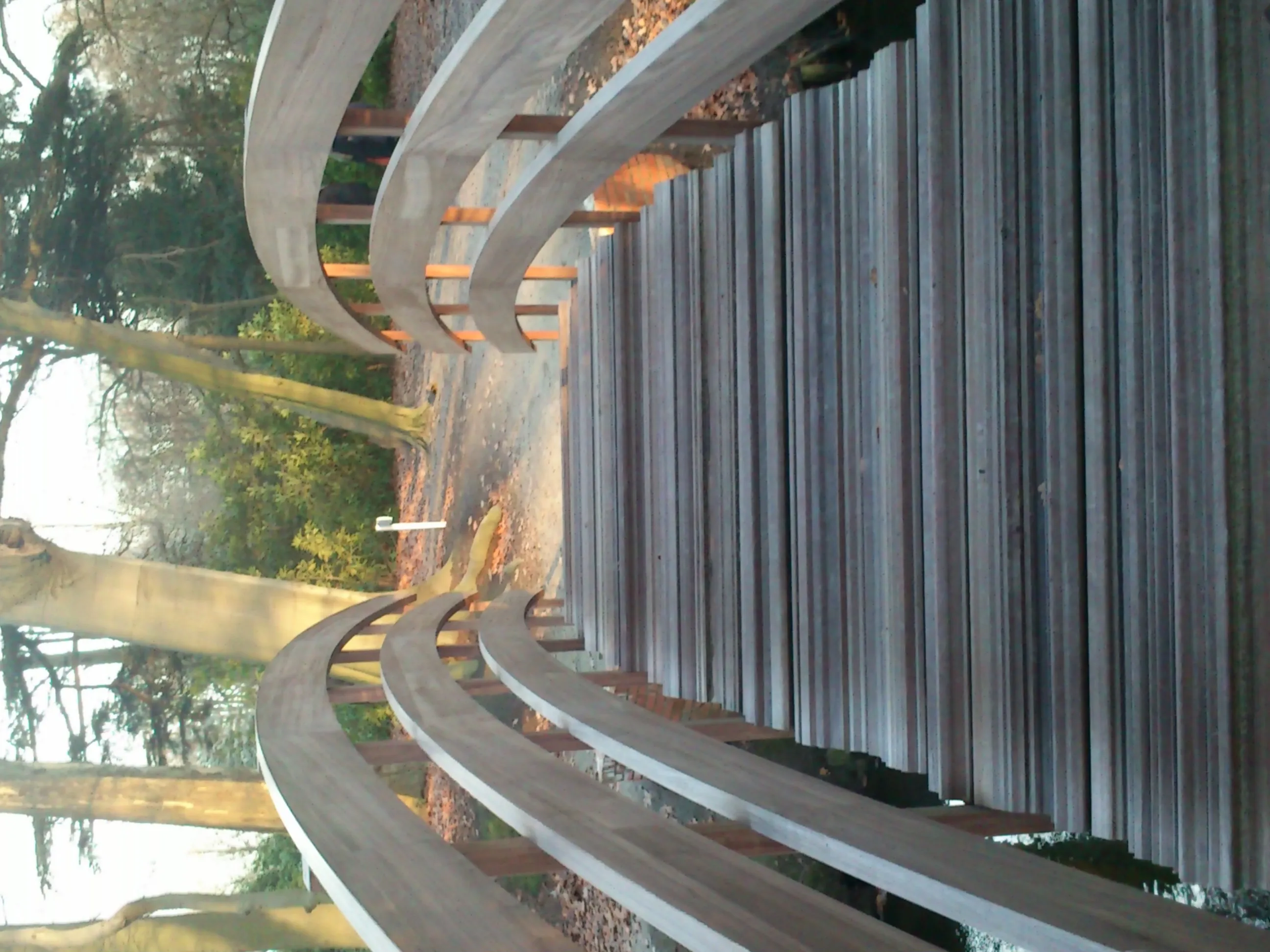 Gran escultura metálica curva con vigas horizontales extendidas y paneles verticales estriados, situada entre árboles en un parque al aire libre.
