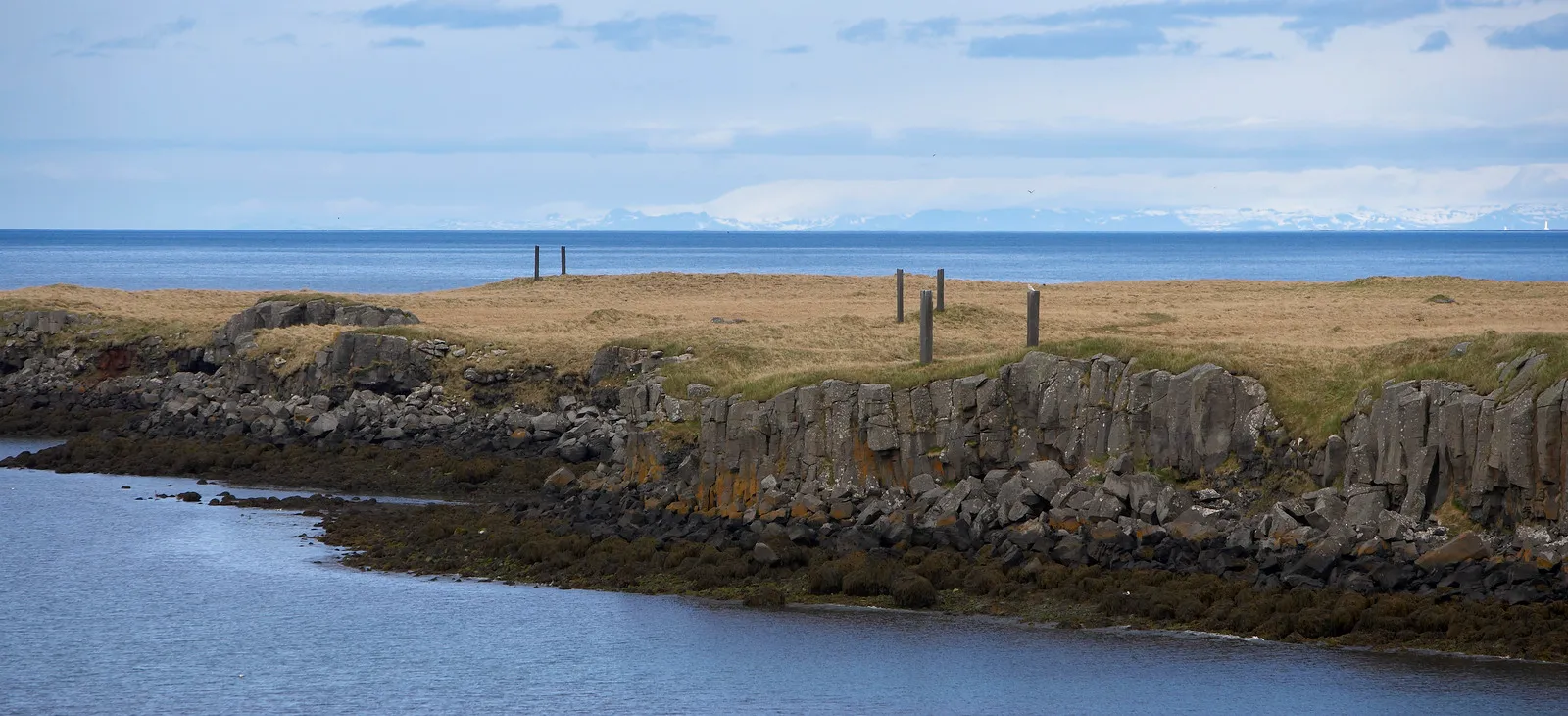 Amplia vista panorámica de la costa de la isla de Viðey, con las columnas de basalto del conjunto escultórico Milestones de Richard Serra sobre la orilla cubierta de hierba frente al horizonte del océano.