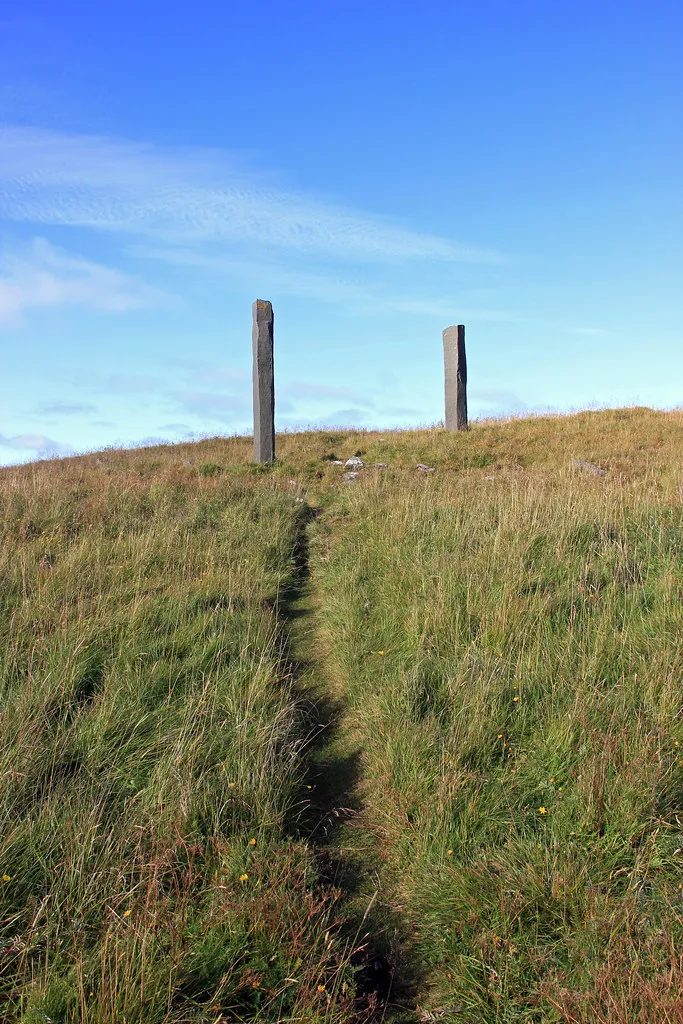 Dos altas columnas de basalto sobre una colina cubierta de hierba bajo un cielo azul, con un sendero estrecho que sube entre la hierba hacia ellas.