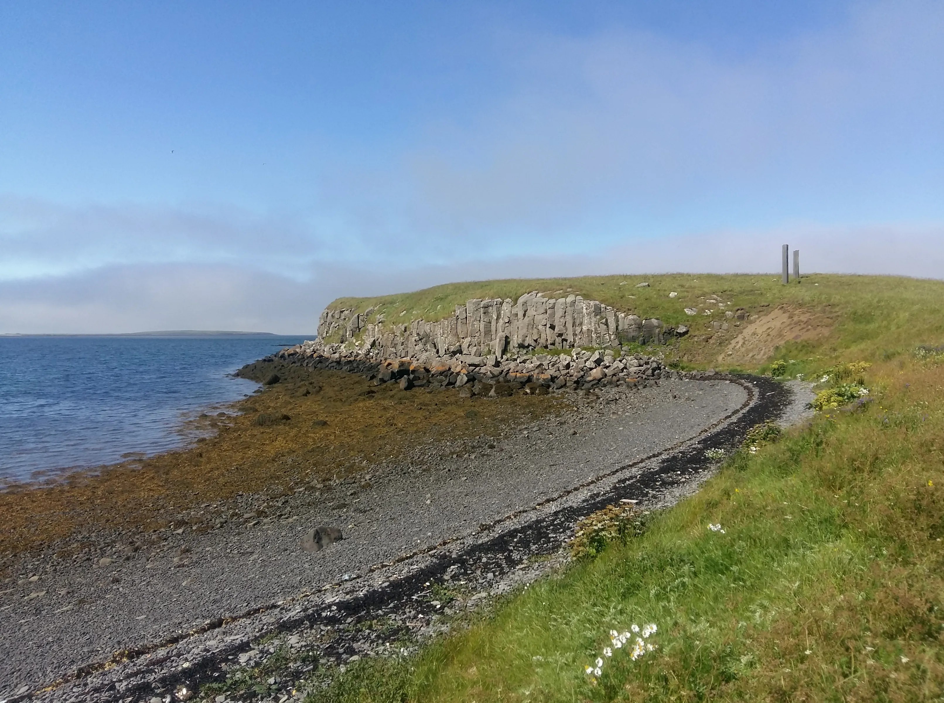 Paisaje costero de la isla de Viðey con una línea de costa rocosa, acantilados cubiertos de hierba y las dos altas columnas de basalto de Milestones de Richard Serra visibles en la cima de la colina bajo un cielo azul