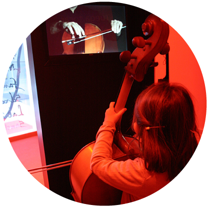 Un niño toca el violonchelo en una habitación iluminada en rojo, frente a una pantalla que muestra un primer plano de unas manos tocando el instrumento con un arco.