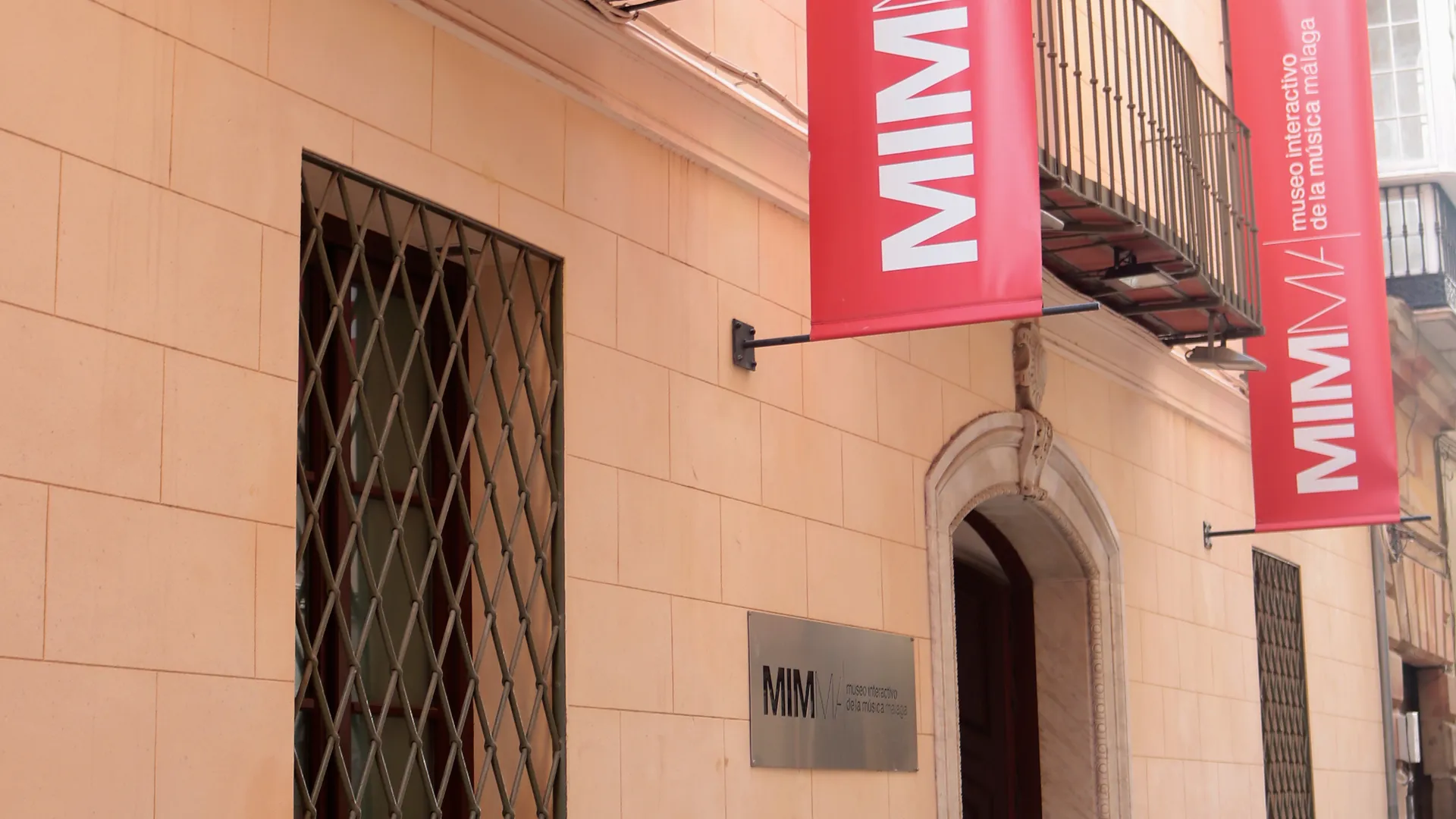 Fachada de piedra beige con pancartas rojas de MIMMA colgadas de un balcón y una pequeña placa de MIM junto a la entrada arqueada