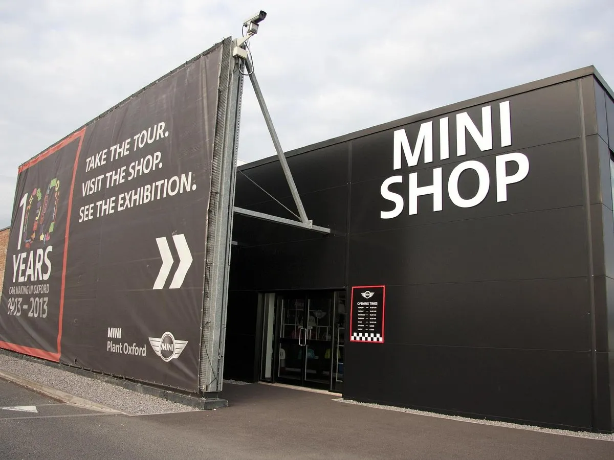 Zewnętrzna część czarnego budynku MINI Shop przy MINI Plant Oxford z dużym białym oznakowaniem oraz banerem reklamującym wycieczki, sklep i wystawy.