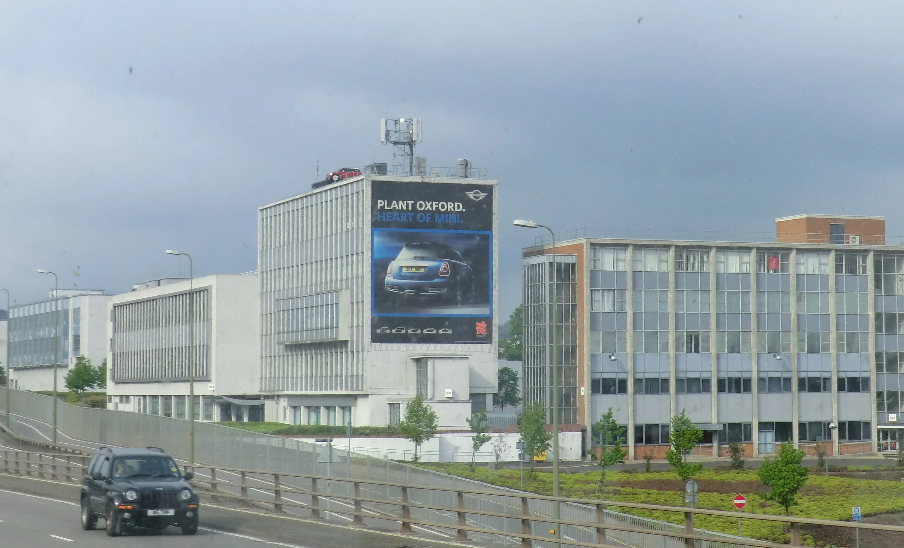 Widok zewnętrzny budynku fabryki MINI w Oksfordzie z dużym billboardem przedstawiającym samochód MINI oraz napisem PLANT OXFORD, z drogą i czarnym samochodem na pierwszym planie.