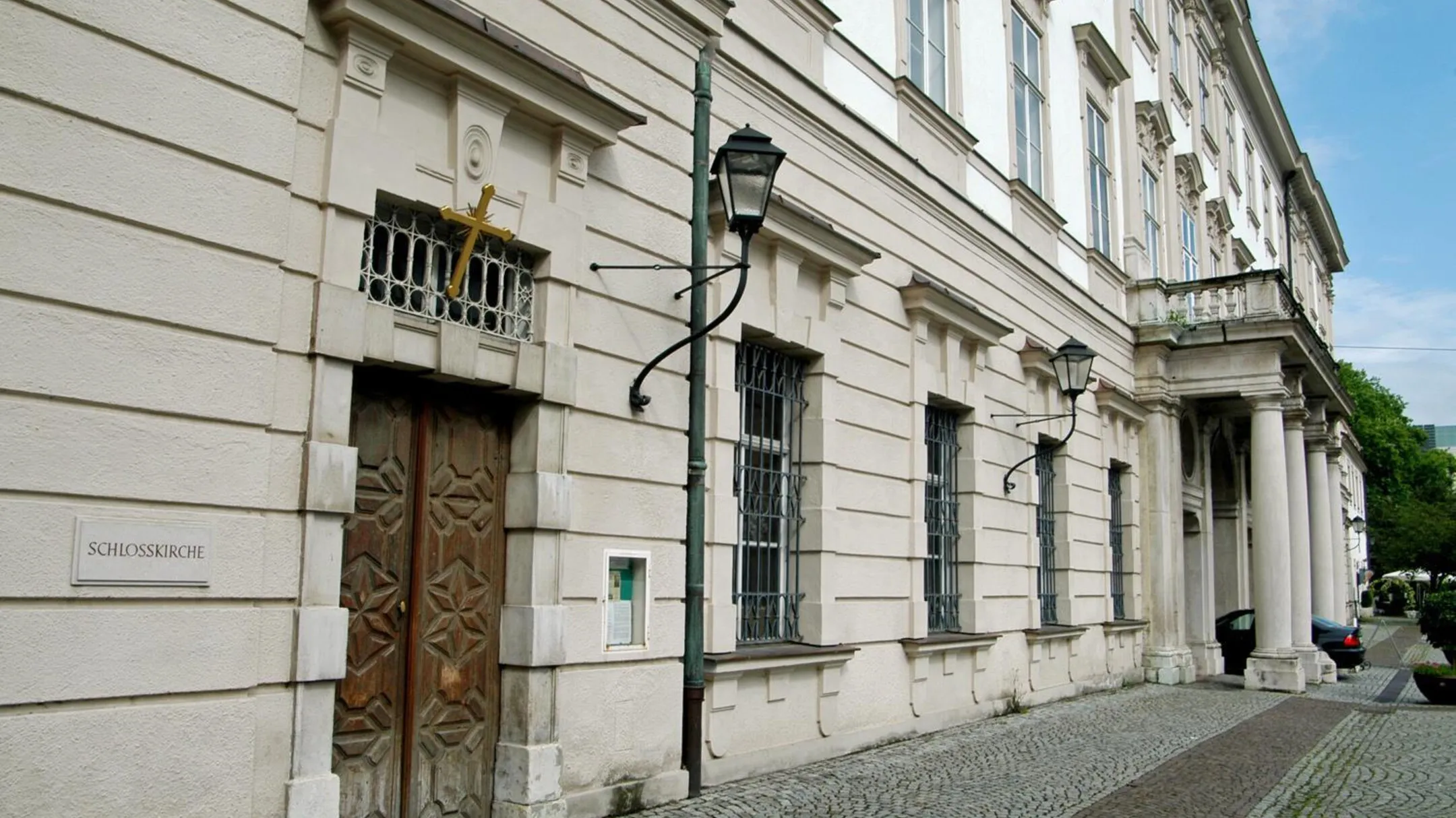 White Baroque building facade with a carved wooden door, iron-grilled windows, a vintage street lamp, and a sign reading Konzertsaal on the wall.