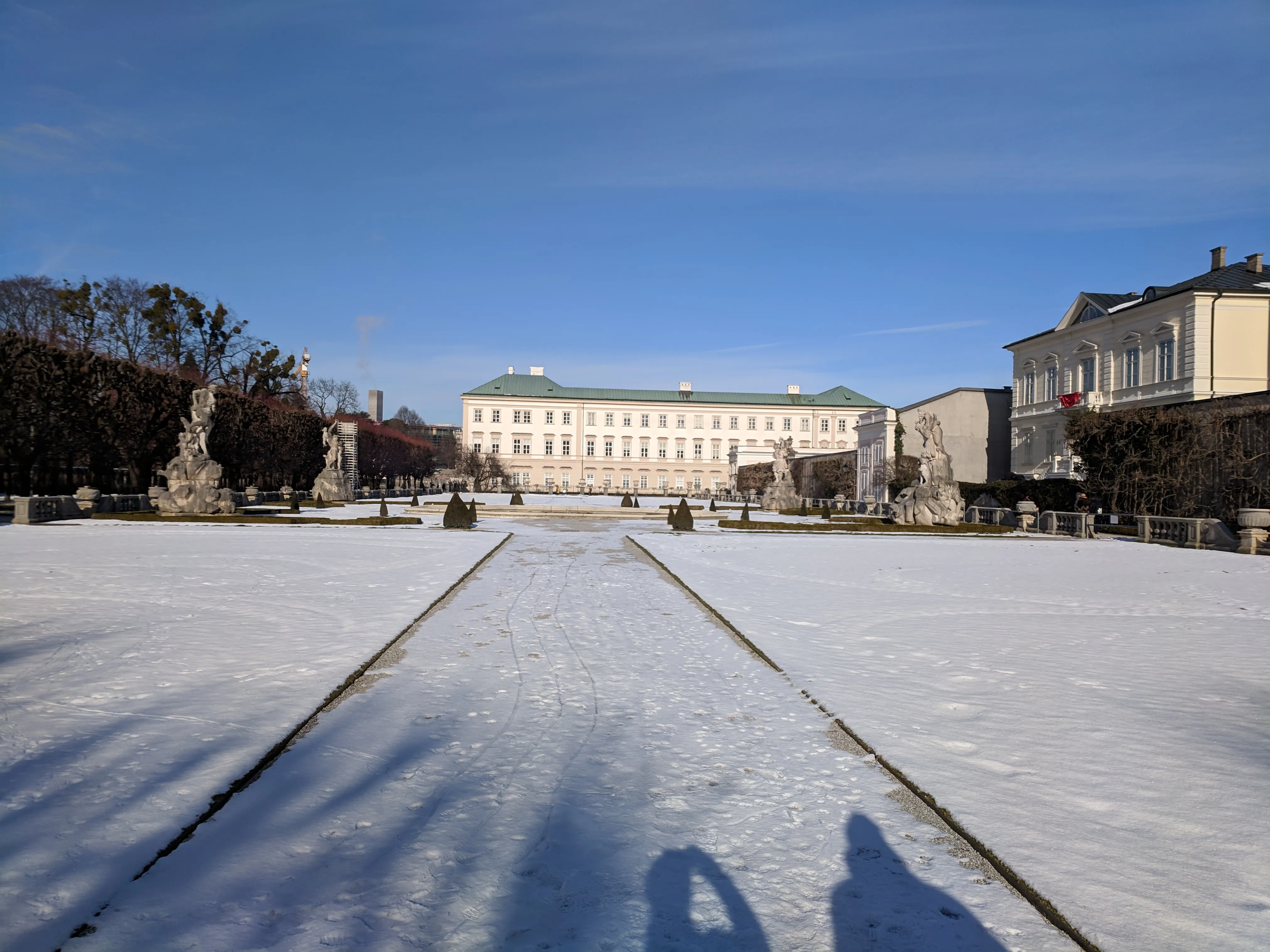 Snow-covered formal garden with a central pathway leading to the white facade of Mirabell Palace, flanked by statues and trimmed hedges under a clear blue sky.