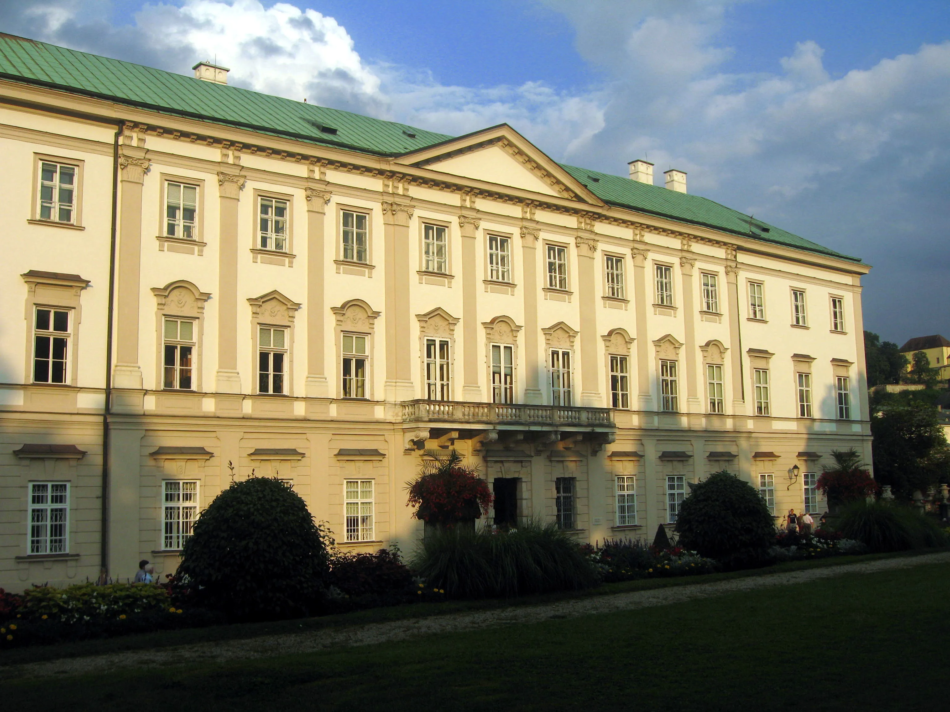 A large baroque palace with cream-colored facade, green roof, and central pediment, viewed from the garden with trimmed bushes and flower beds in the foreground
