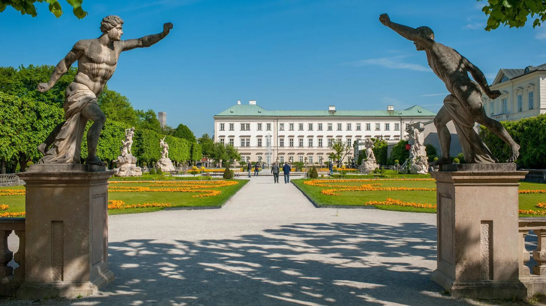 Two large statues on pedestals frame a central garden pathway leading to the white baroque Mirabell Palace, with orange flower beds and green lawns on either side under a clear blue sky.