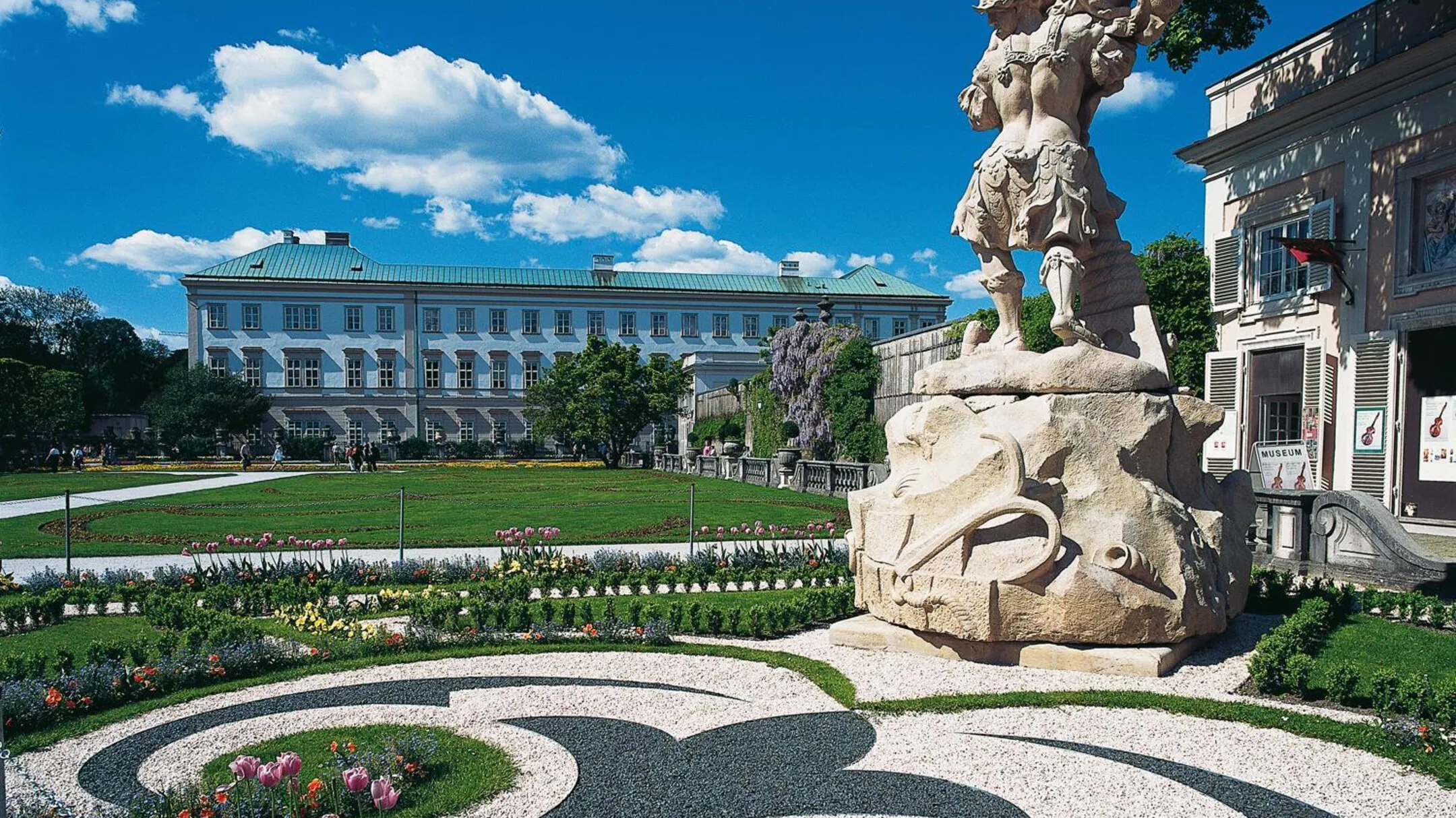 Wide view of Mirabell Palace gardens with a large baroque stone sculpture in the foreground, formal parterre flower beds with gravel pathways, and the white palace building under a blue sky with clouds.