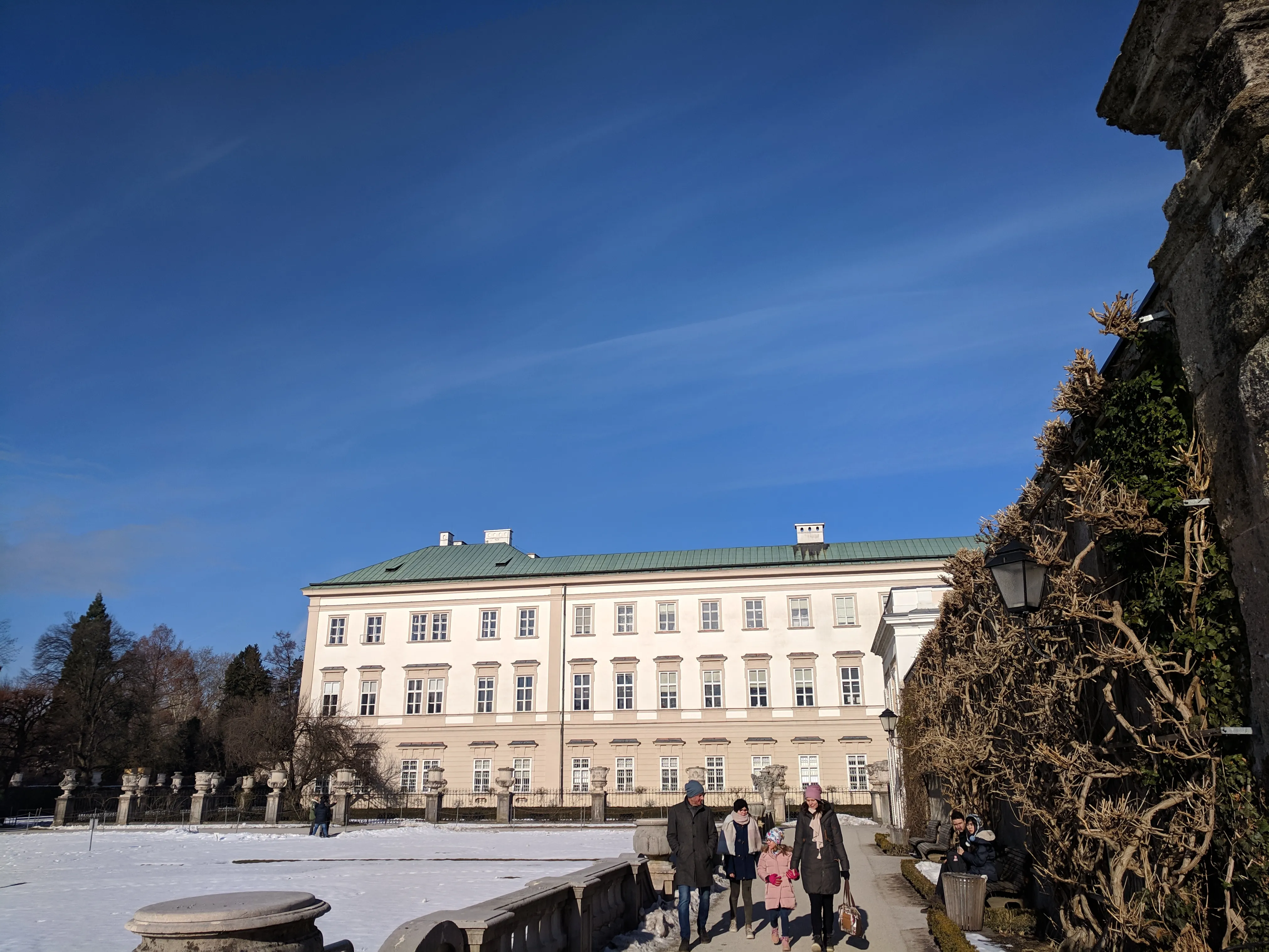 White baroque Mirabell Palace with green roof under a clear blue sky, with snow on the ground and people walking along a path beside a stone wall