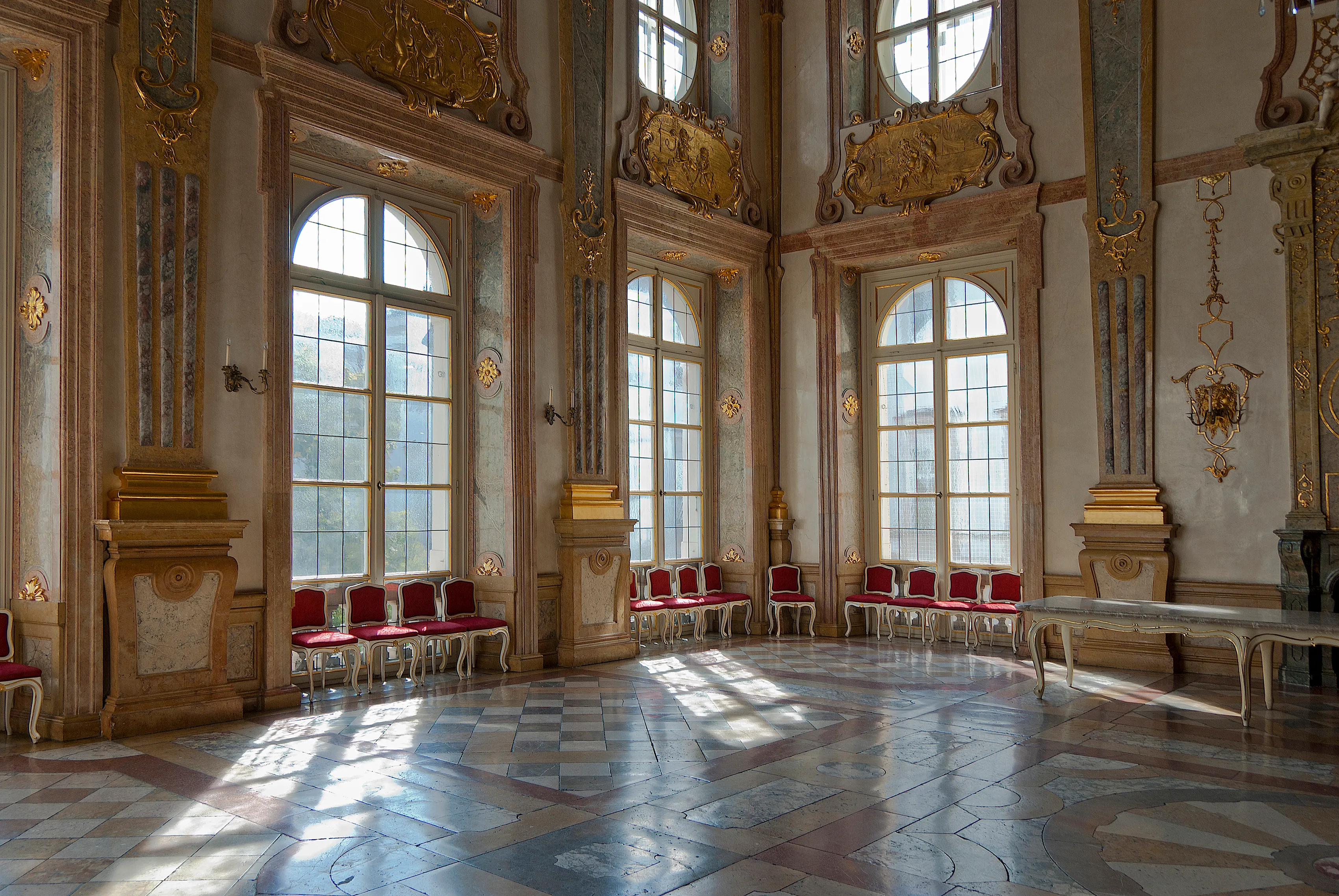 Ornate baroque hall interior with tall arched windows, gilded stucco decorations, red velvet chairs lined along the walls, and a checkered marble floor with sunlight streaming in.
