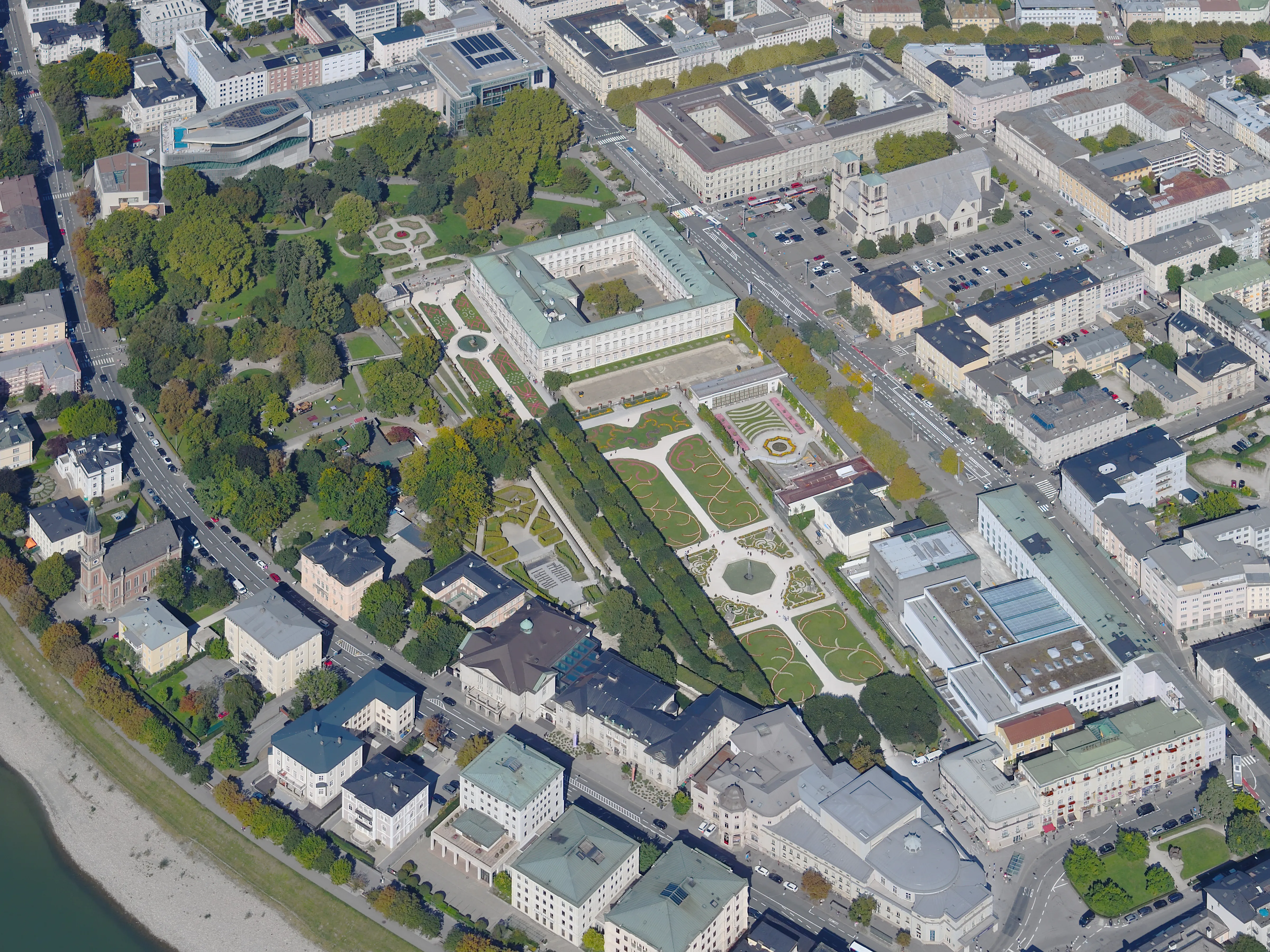 Aerial view of Mirabell Palace and its formal baroque gardens with geometric parterres, surrounded by city buildings and the Salzach River along the left edge.