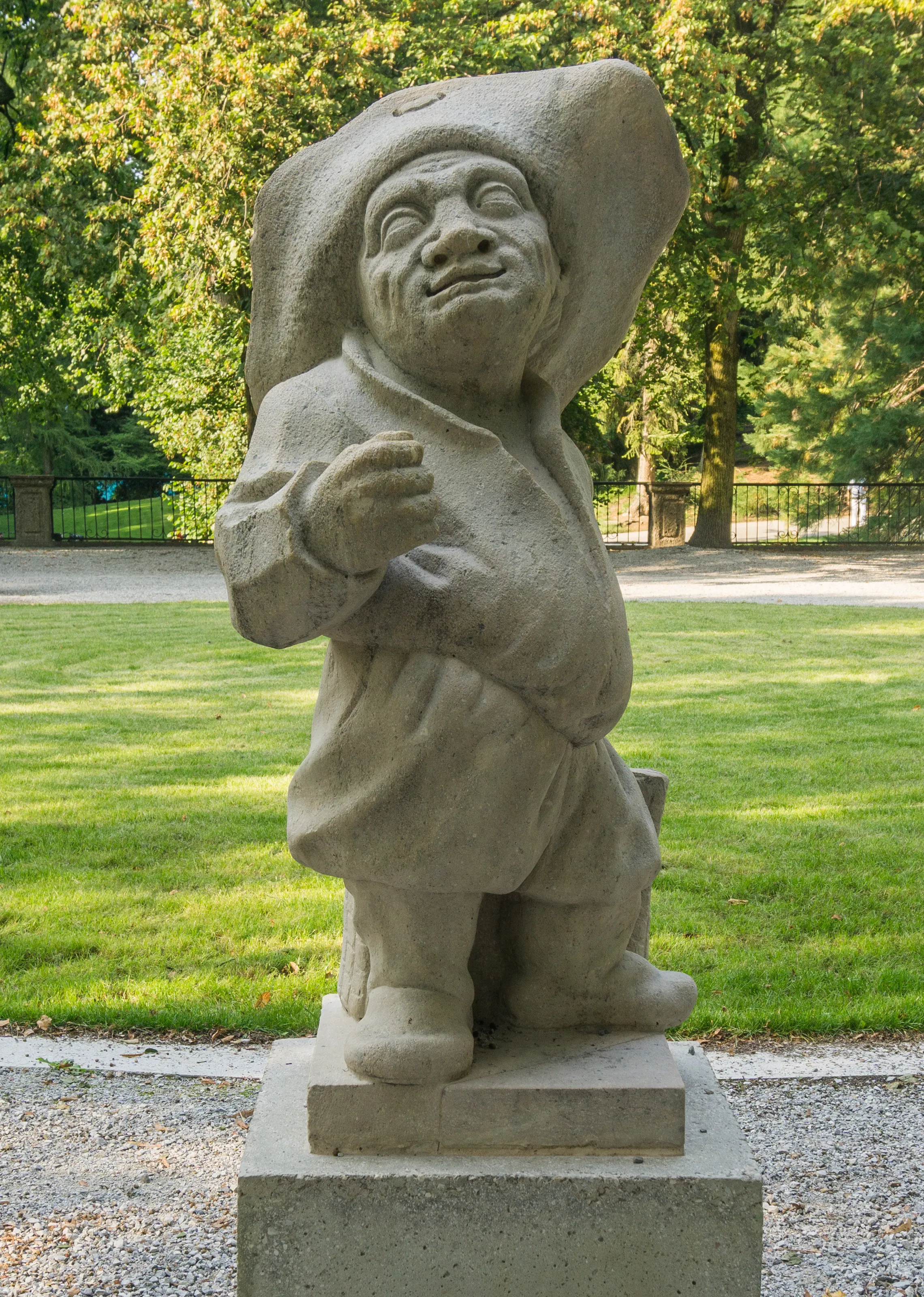 Stone statue of a smiling dwarf figure wearing a large hat, standing on a pedestal in a grassy garden with trees in the background.