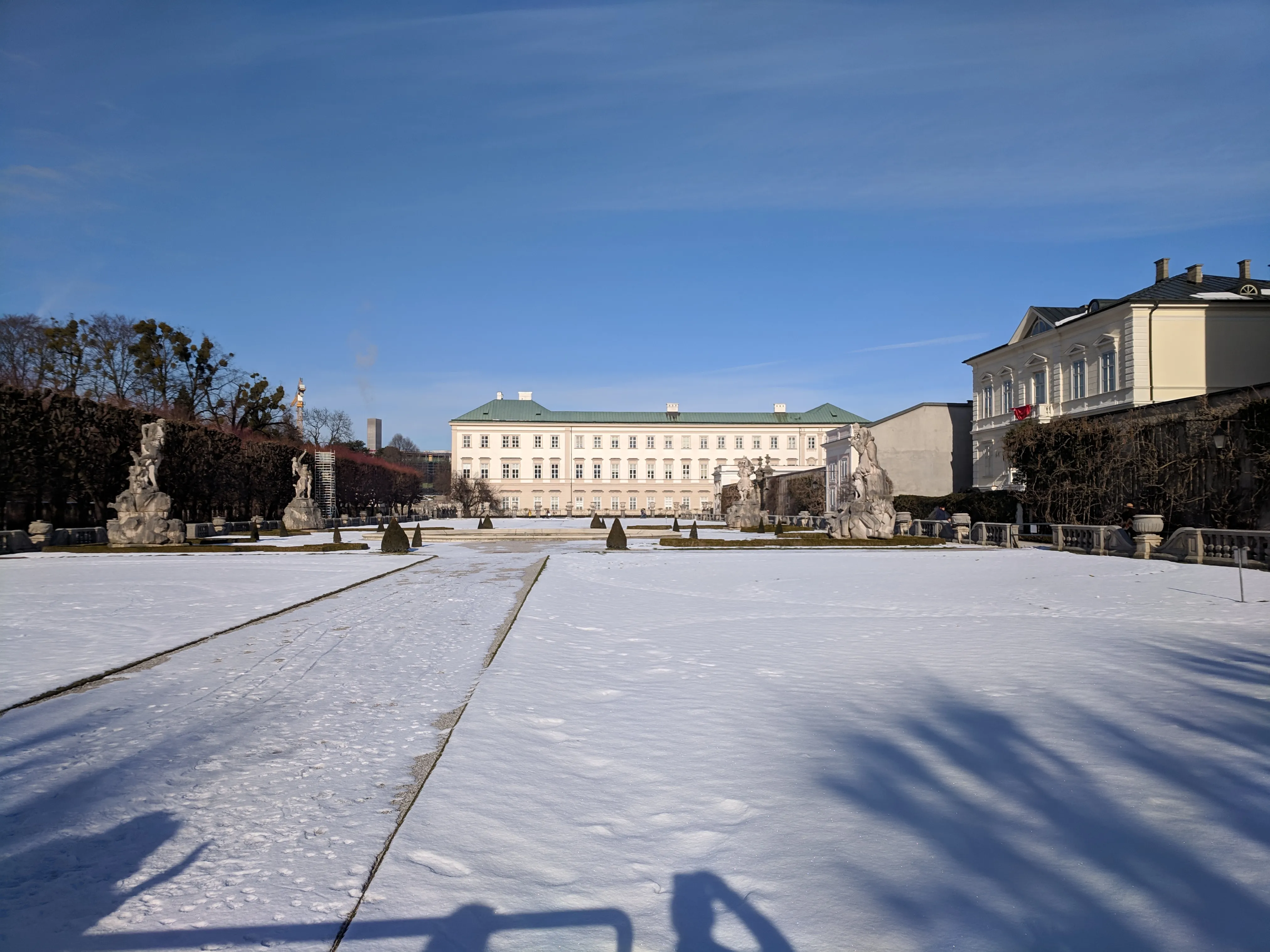 Snow-covered formal gardens leading to the pale facade of Mirabell Palace under a clear blue sky, with statues and trimmed hedges lining the path.