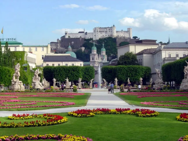 Formal garden with colorful flower beds, a central path leading to a fountain, flanked by statues and hedges, with a large fortress on a hill in the background.