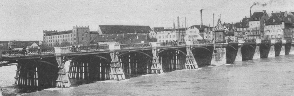 Historische zwart-witfoto van de stenen boogbrug Mittlere Brücke over de Rijn, met houten steigers en stadsgebouwen waaronder de Käppelijoch-kapel op de achtergrond