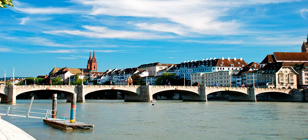 Wijd panoramisch uitzicht op de stenen boogbrug Mittlere Brücke over de Rijn in Basel, met de skyline van de stad en de toren van de Basler Münster op de achtergrond onder een blauwe lucht.