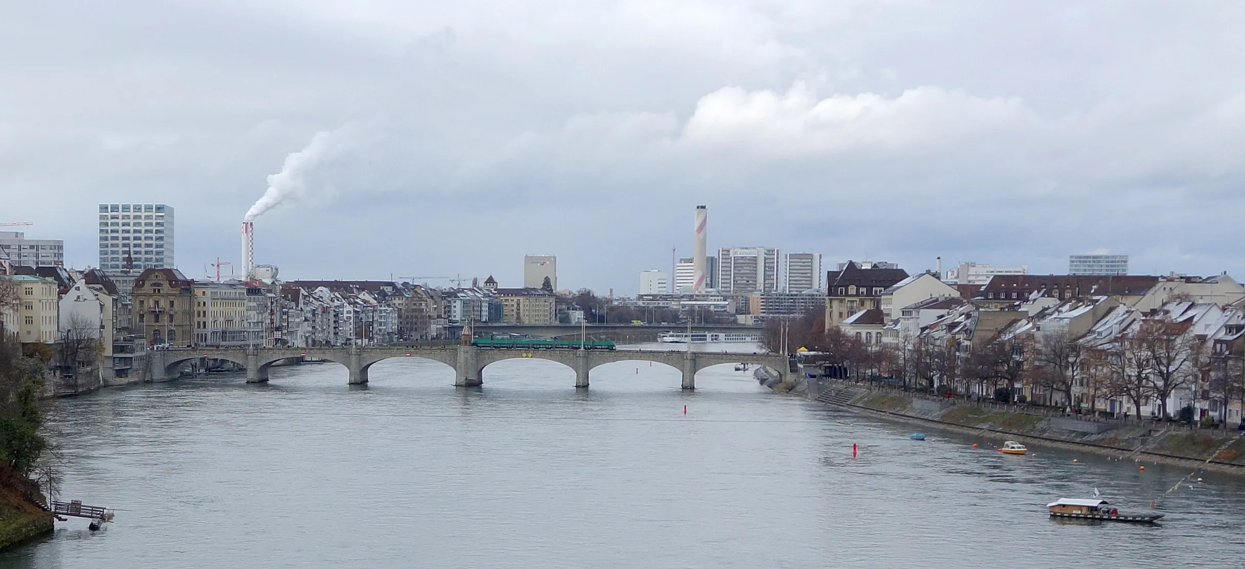 Breed panoramisch uitzicht op de stenen boogbrug Mittlere Brücke over de Rijn in Bazel, met stadspanden aan beide oevers en een schoorsteen die witte rook uitstoot op de achtergrond onder een bewolkte hemel.