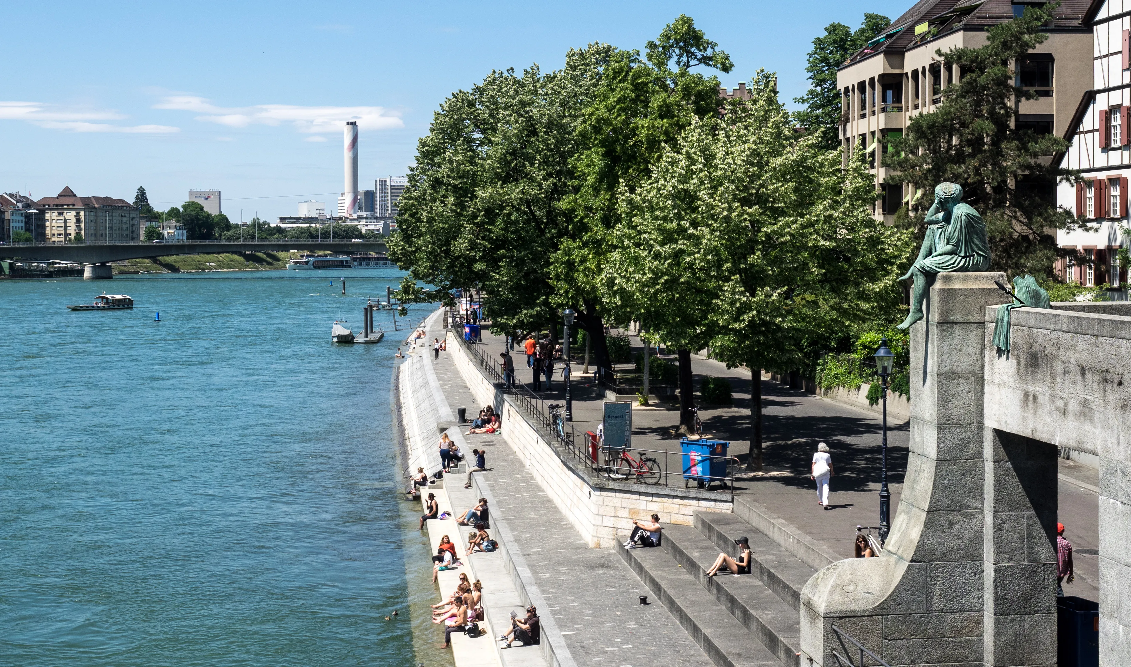 Mensen die op de trappen langs de oever van de Rijn in Basel zitten, met de stenen pijler en het bronzen standbeeld van de Mittlere Brücke zichtbaar aan de rechterkant, bomen langs de promenade en een brug die de rivier in de achtergrond oversteekt.