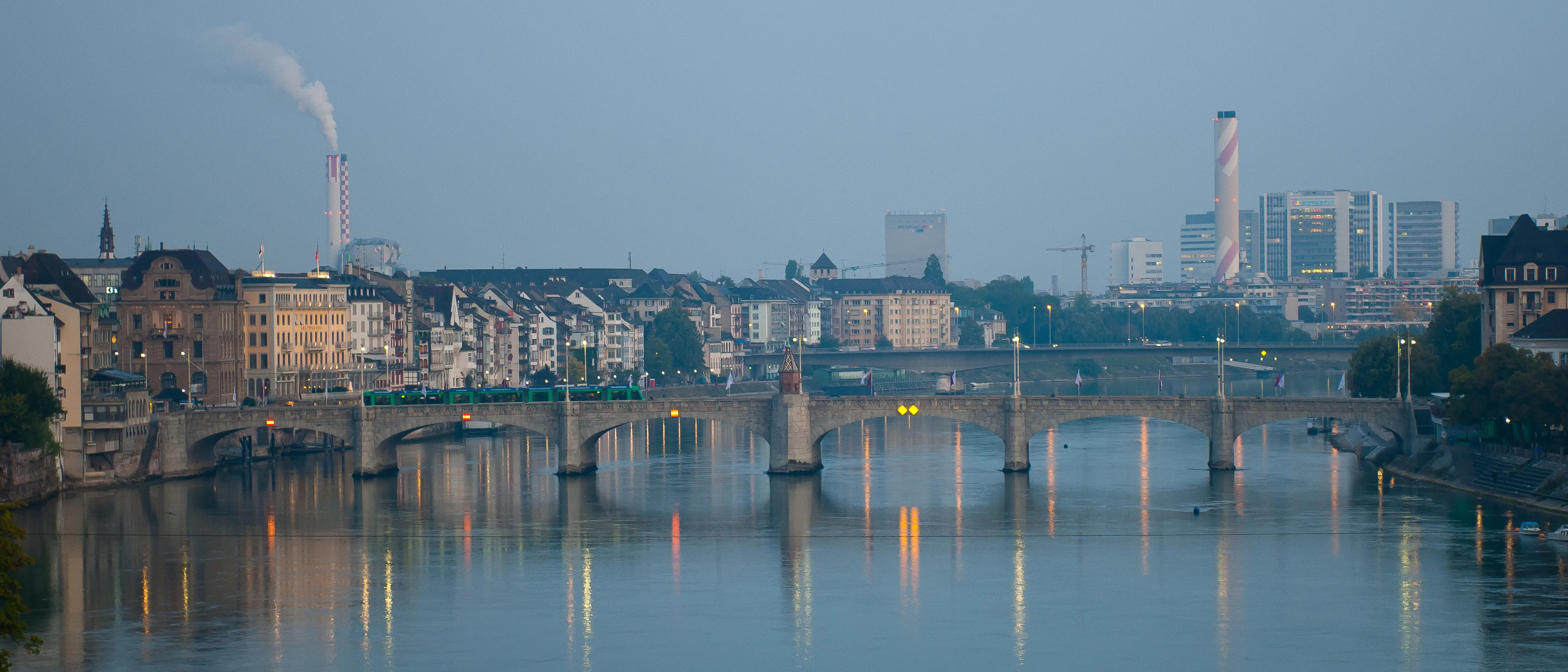 Breed panoramisch uitzicht op de stenen boogbrug Mittlere Brücke over de Rijn in Bazel bij schemering, met stadsgebouwen en twee hoge industriële schoorstenen die weerspiegelen in het kalme water.