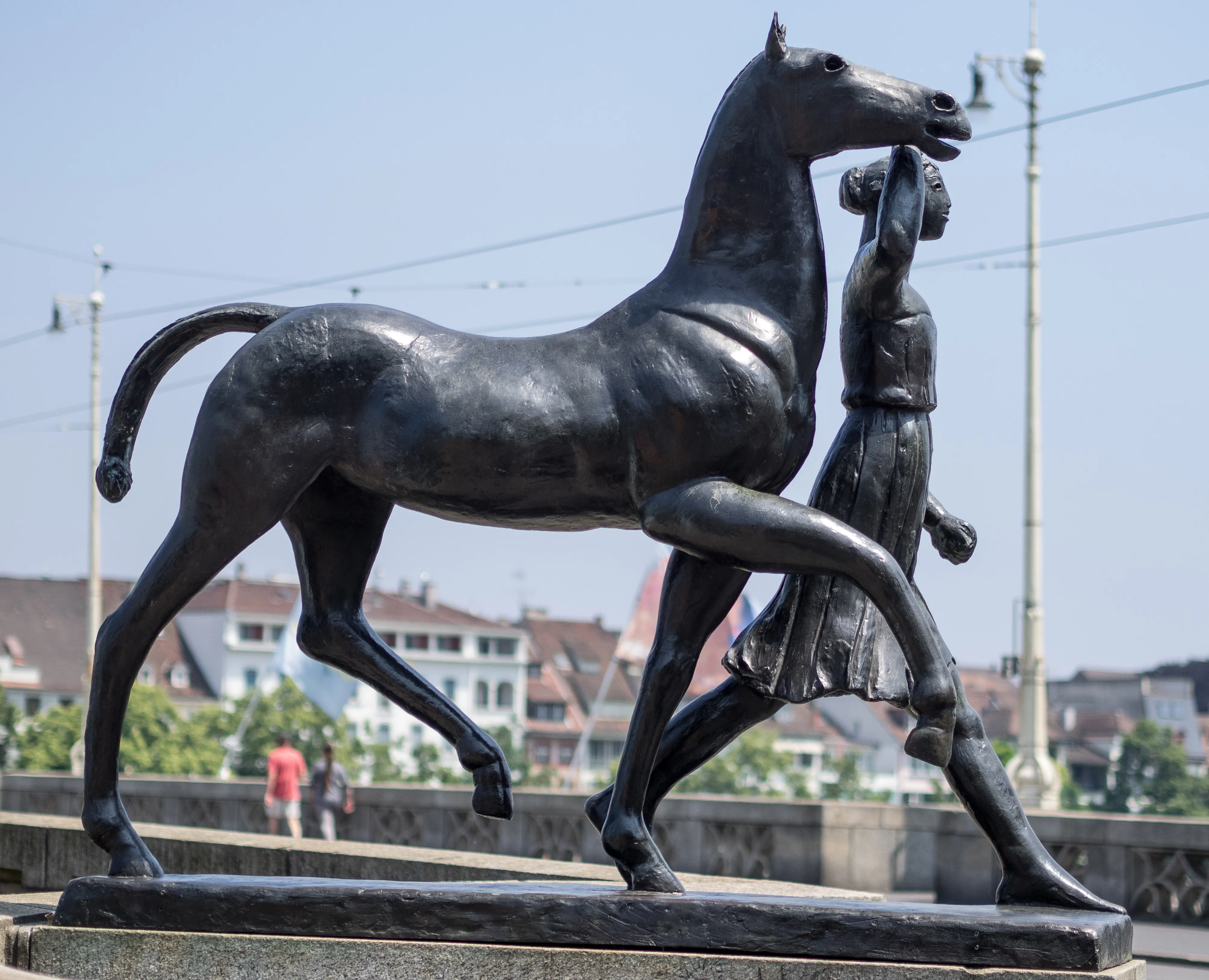Bronzen sculptuur van een paard en een vrouwenfiguur op een stenen voetstuk op een brug, met stadsgebouwen en een blauwe lucht op de achtergrond