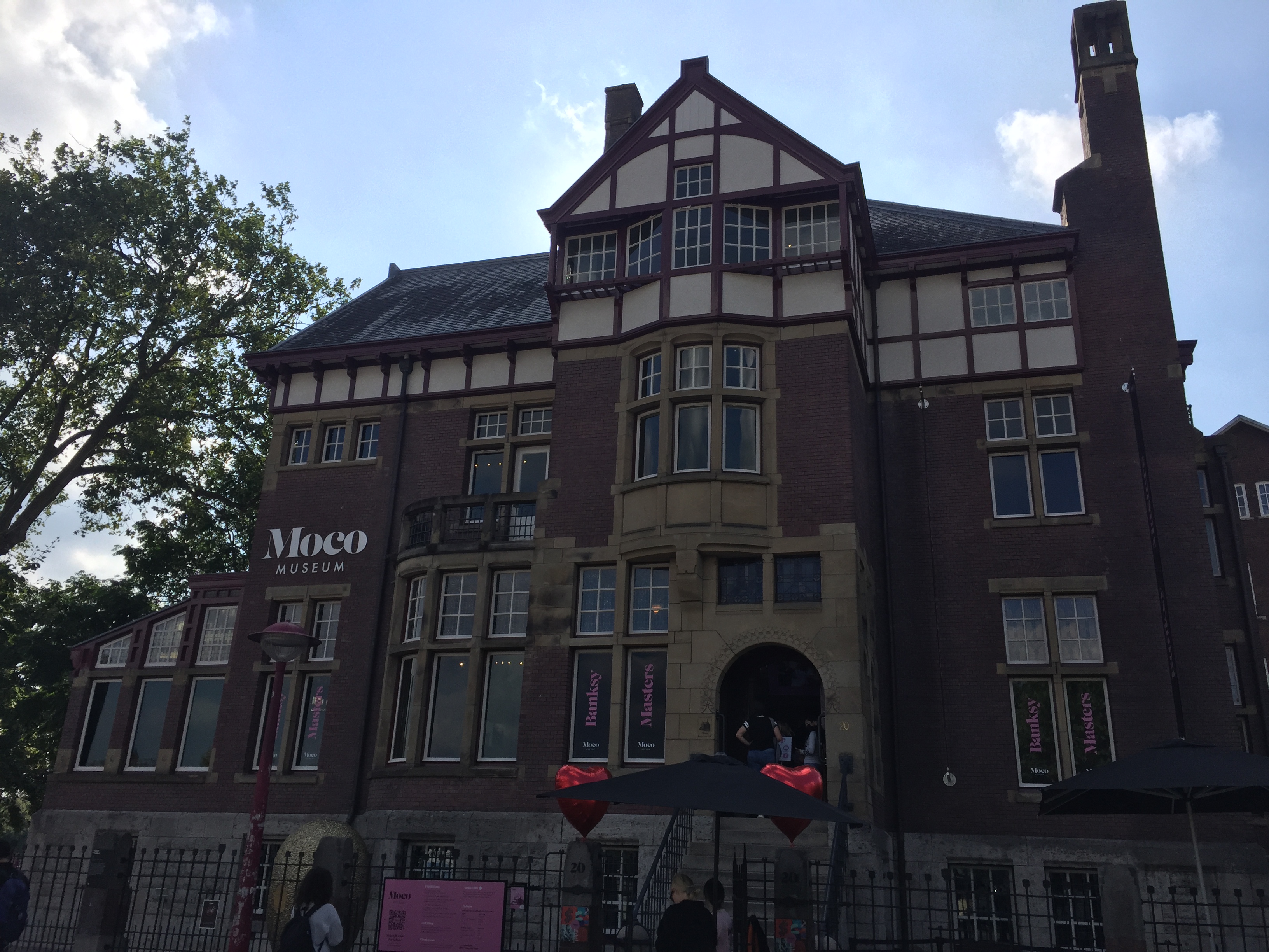 Exterior del edificio del Museo Moco, una villa histórica con ladrillos de color rojo oscuro, entramado de madera blanco y un arco de entrada prominente bajo un cielo azul