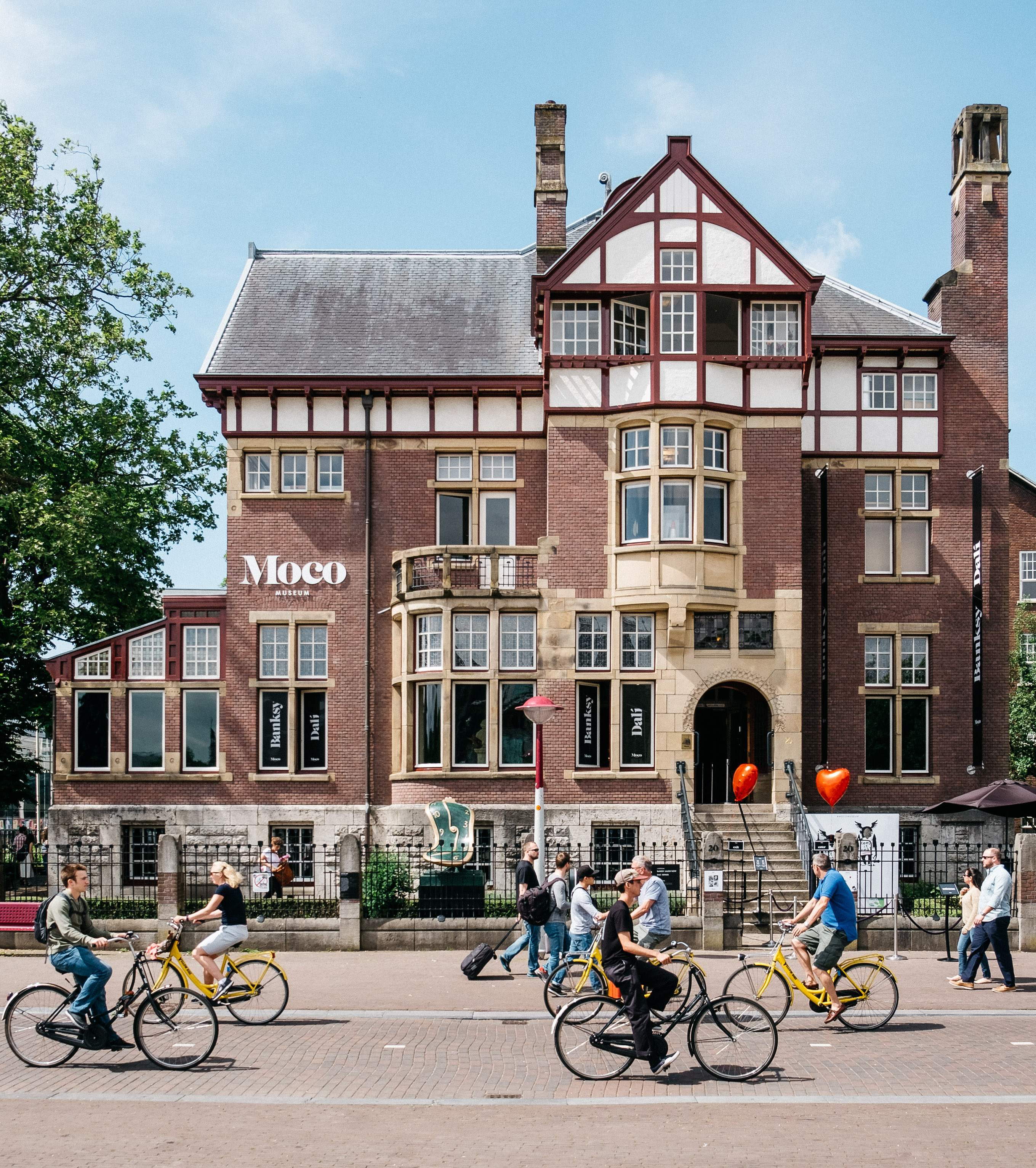 Edificio histórico de ladrillo rojo con arquitectura Tudor que muestra el letrero 'Moco', gente andando en bicicleta en la calle de enfrente bajo un cielo azul.
