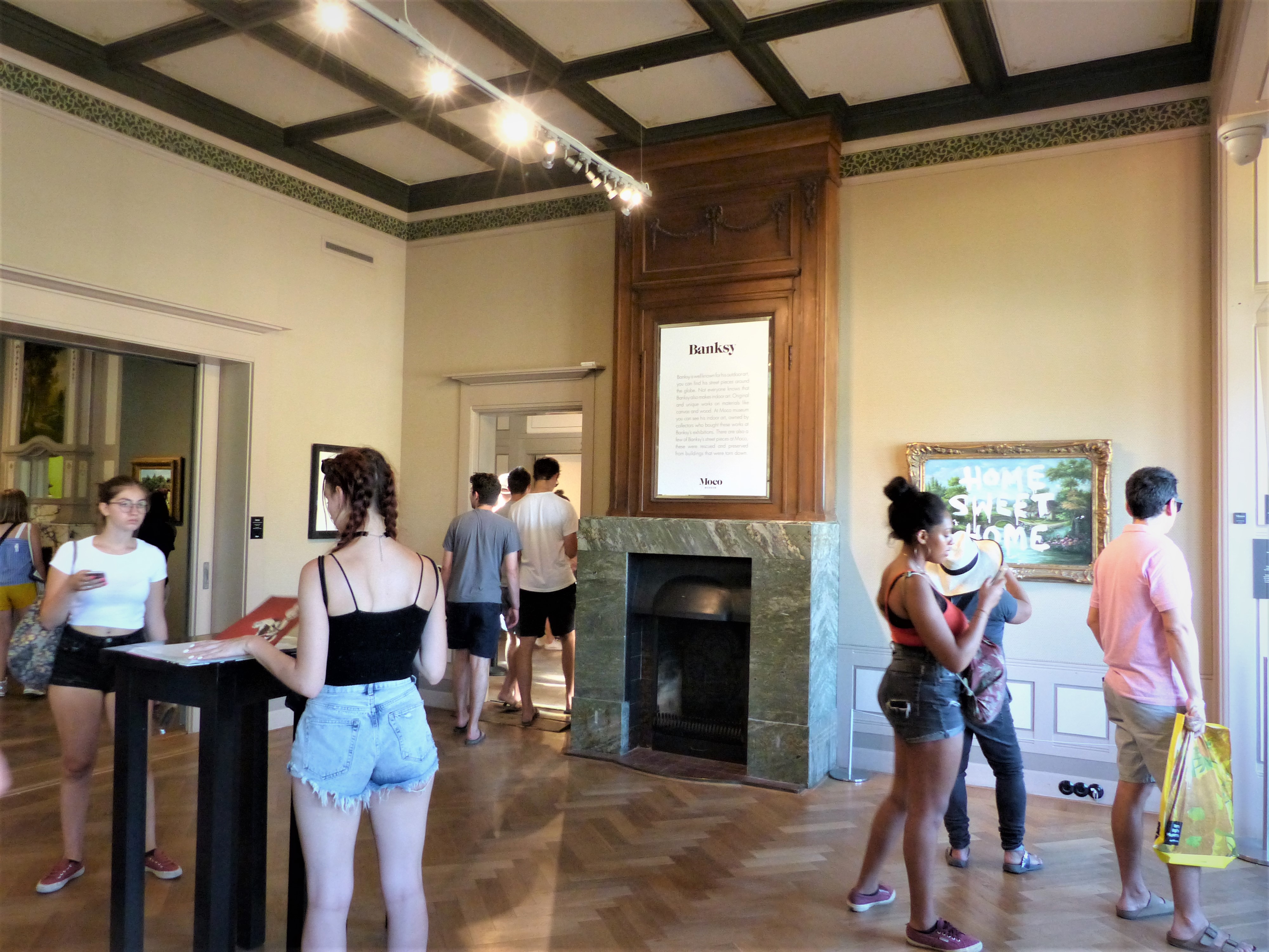 Amplio interior de museo con visitantes cerca de una chimenea de mármol y obras de arte enmarcadas en la pared bajo vigas expuestas en el techo