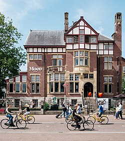 El edificio del Museo Moco, una villa histórica de ladrillo rojo con detalles blancos, con el letrero 'Moco', rodeado de gente en bicicleta y caminando en un día soleado.