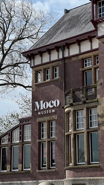 Fachada de edificio de ladrillo con letrero de 'Moco Museum', ventanas altas, molduras de piedra y árboles sin hojas bajo un cielo parcialmente nublado