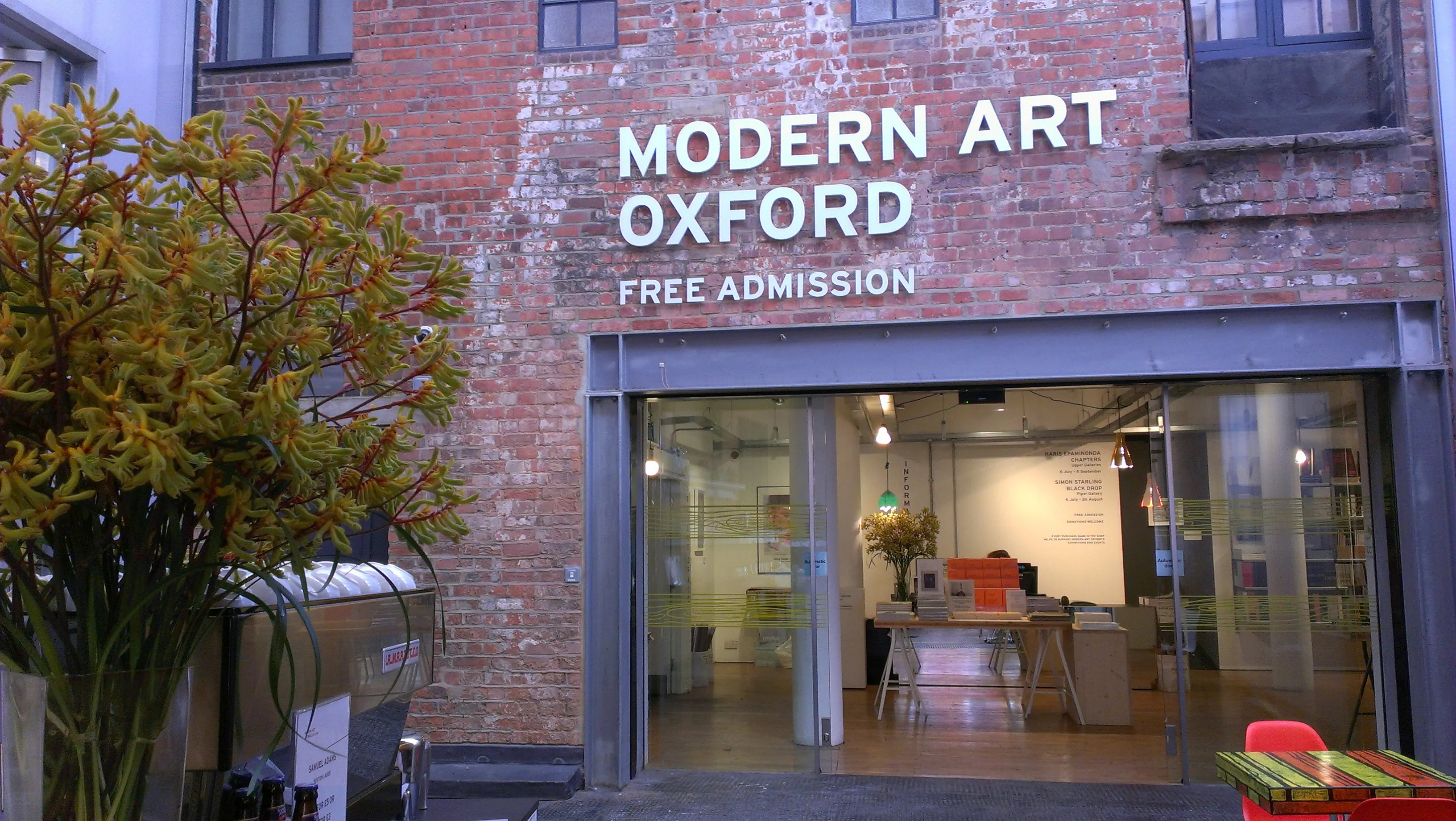 Red brick building facade with 'MODERN ART OXFORD' and 'FREE ADMISSION' white lettering above a glass entrance doorway revealing an interior lobby