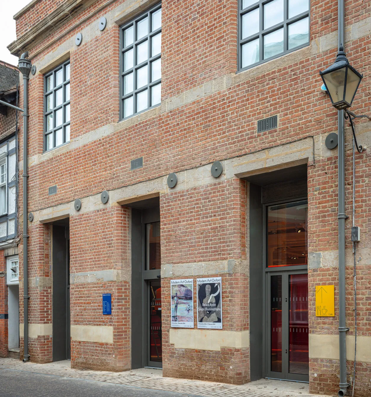 Red brick building facade with three dark-framed entrances, large grid windows on the upper floor, exhibition posters on the wall, and a street lamp